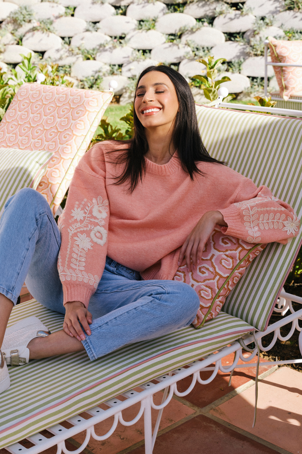 A woman sits smiling on a striped outdoor lounge chair, wearing a dusty pink knit sweater with white floral patterns on the sleeves and light blue jeans.