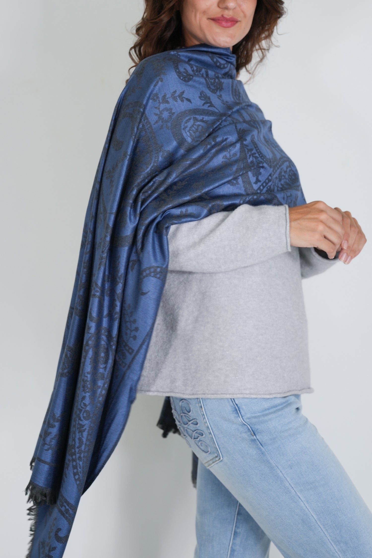 Woman wearing a blue patterned shawl over a gray sweater and light blue jeans on a white background