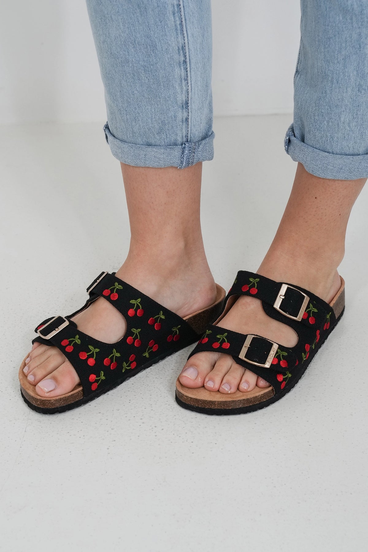 Black double-strap sandals with a red and green cherry pattern and gold-tone buckles, shown on a model wearing light blue cuffed jeans.