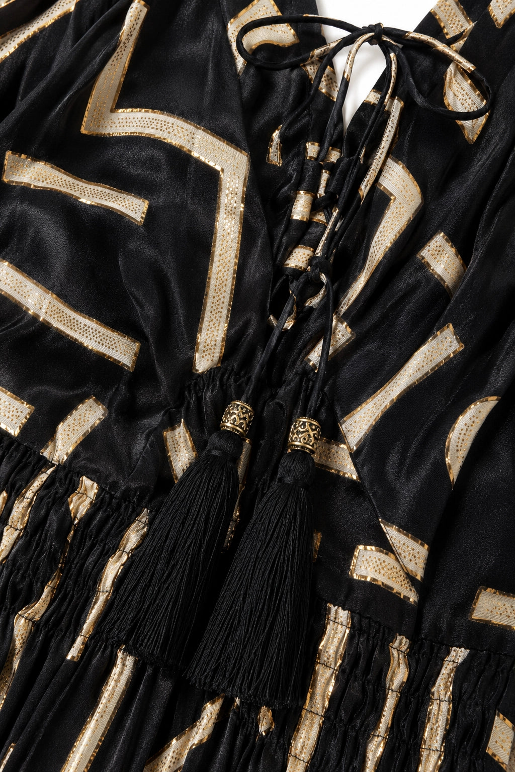 Close-up of a black velvet garment with metallic gold geometric stripes and black tassels tied at the neckline.