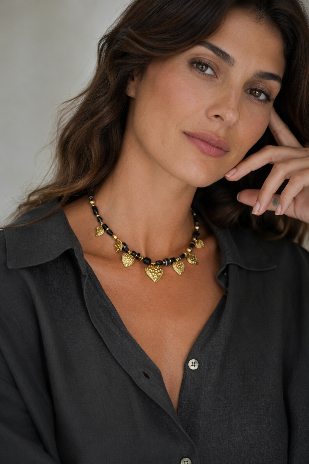 A close-up of a woman wearing a gold heart-shaped pendant necklace with black and gold beads, paired with a dark gray button-up shirt.