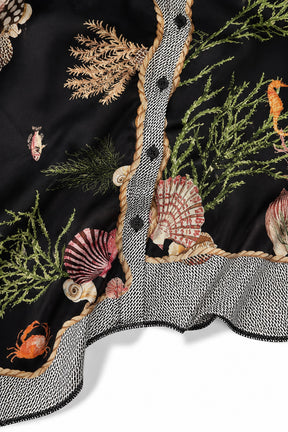 Close-up of a black embroidered fabric with an undersea theme, featuring seashells, coral, seaweed, and small sea life motifs, finished with a gray and white patterned ruffled trim and black buttons.