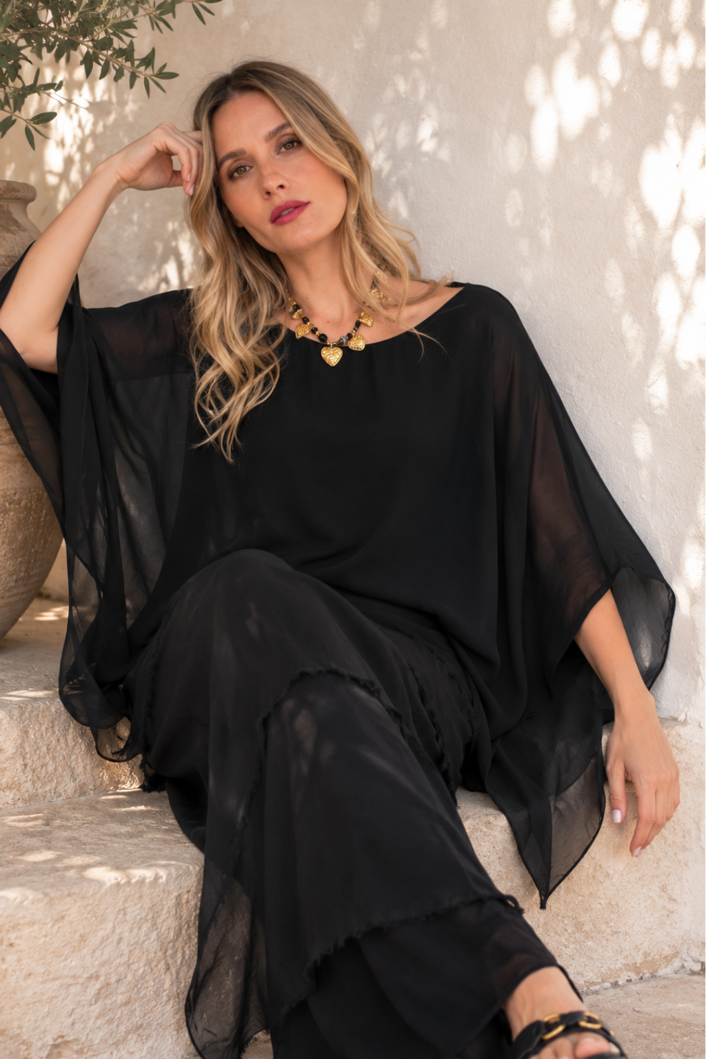 A woman wearing a black sheer long-sleeve maxi dress sits on stone steps against a textured wall with dappled sunlight.