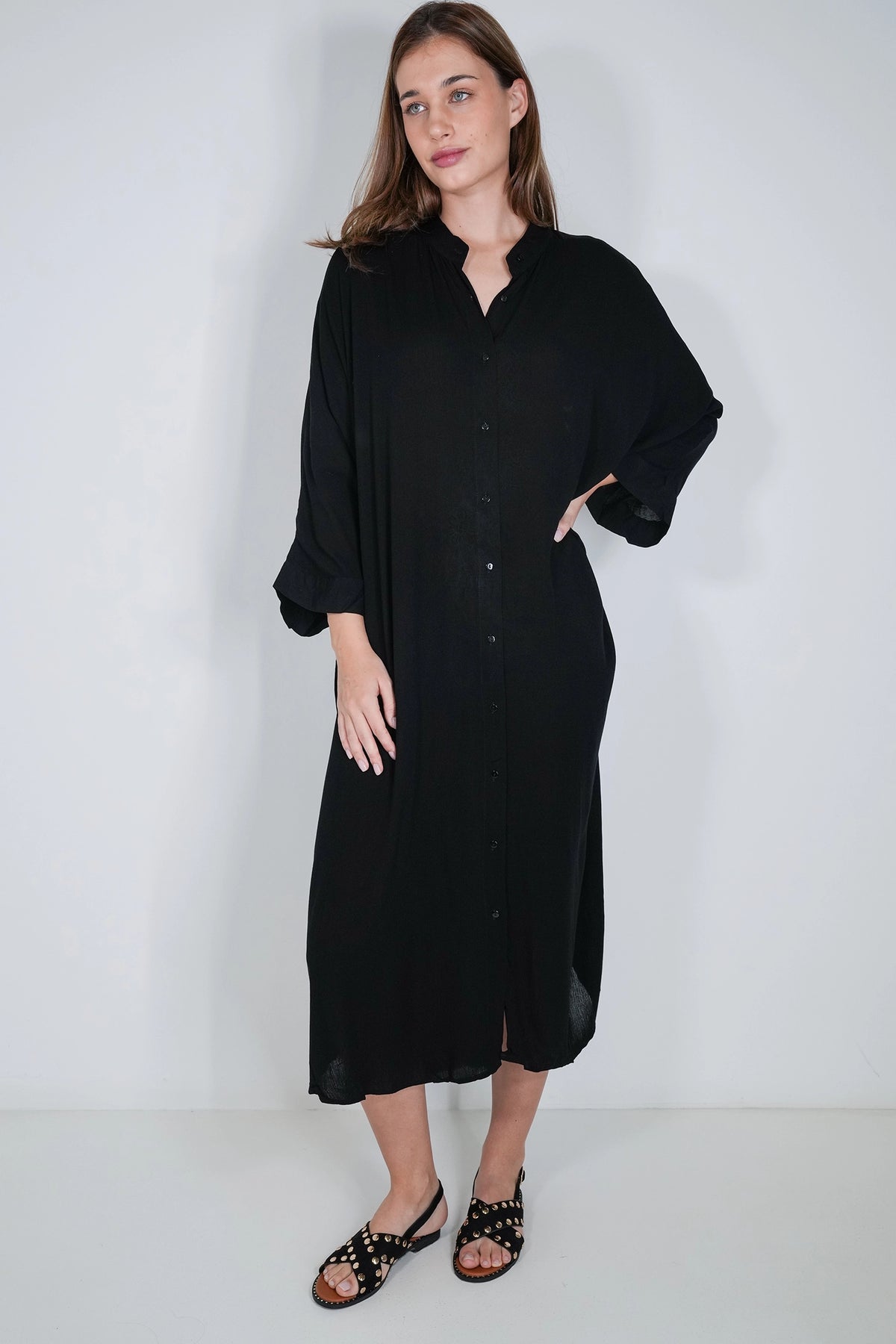 A woman modeling a loose black midi shirt dress with three-quarter sleeves and a button-down front, paired with black studded sandals, against a plain light gray studio background.