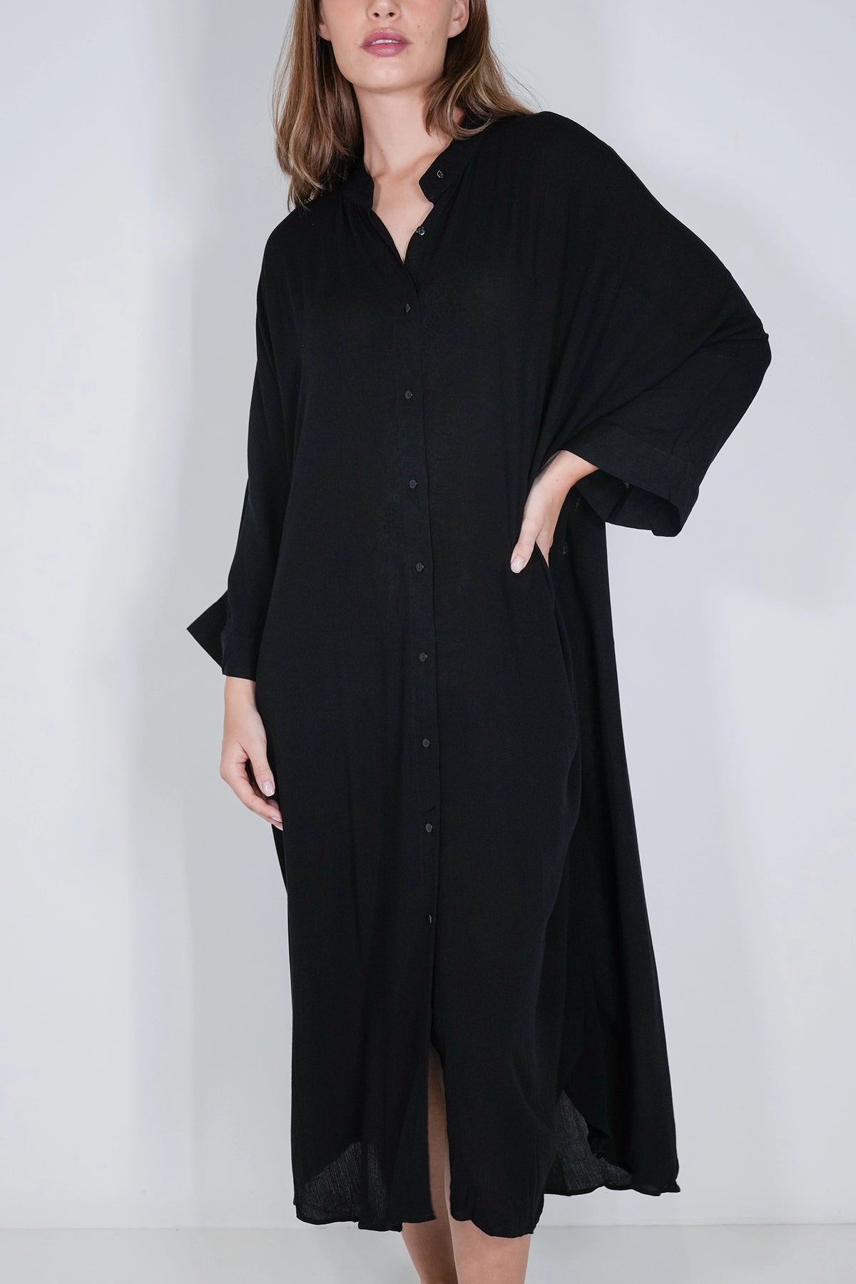 A woman wearing a loose, long black button-front dress or tunic with wide draped sleeves, photographed against a plain light gray studio background.