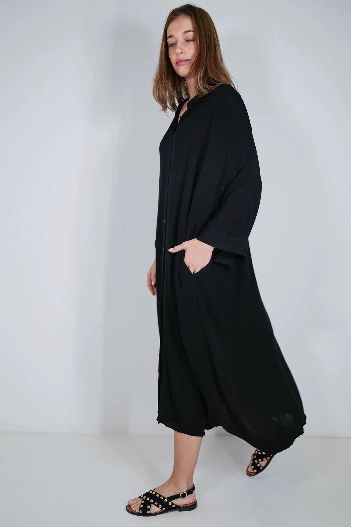 A woman modeling a long black button-front abaya dress with wide sleeves and side pockets, paired with black studded sandals, against a plain light gray studio background.