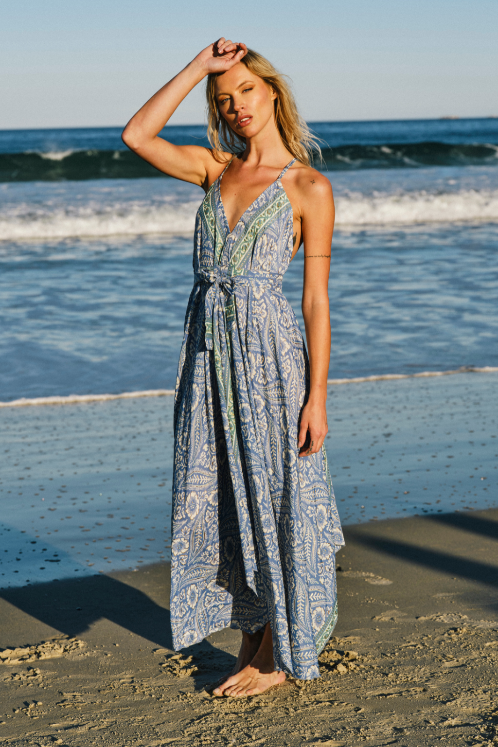 A woman stands barefoot on a sandy beach wearing a long, light blue maxi dress with a green and white paisley-style print. The dress has a wrap-style bodice with thin straps and a tied waist.
