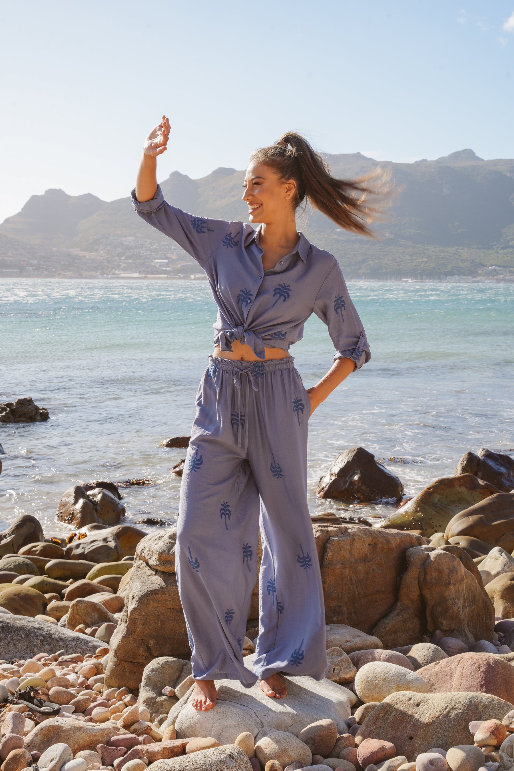 Woman in a light blue outfit standing on a rocky beach with mountains in the background