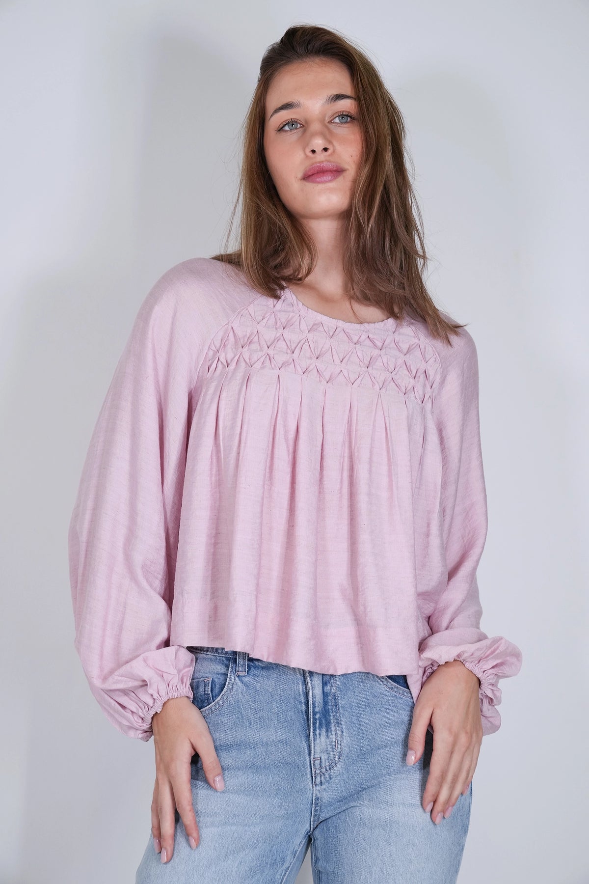 A woman wearing a light pink long-sleeve blouse with a smocked yoke and gathered cuffs, paired with light blue jeans, standing against a plain light gray background.