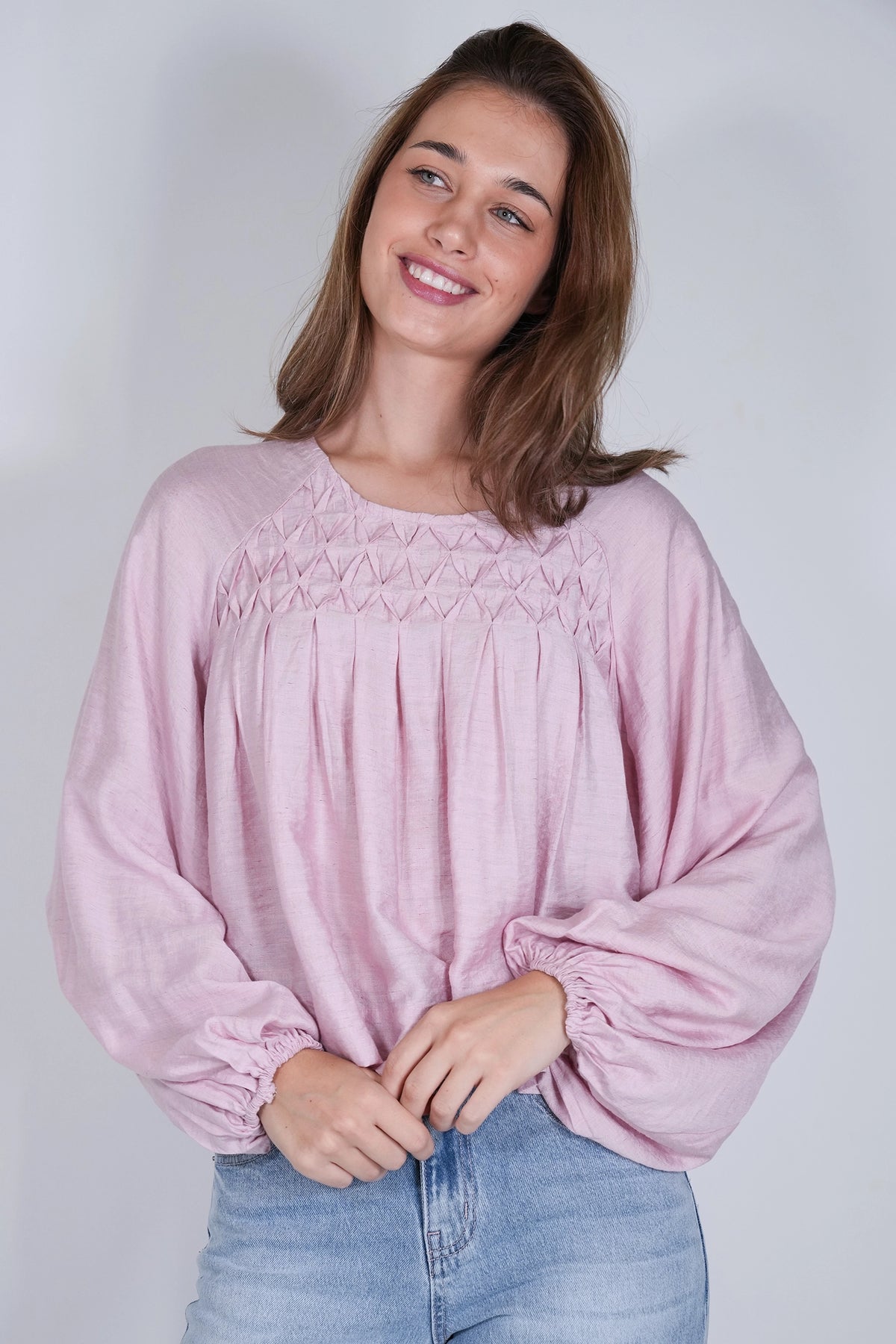 A woman wearing a dusty pink long-sleeve blouse with a smocked chest panel and gathered cuffs, paired with light blue jeans, against a plain light gray background.