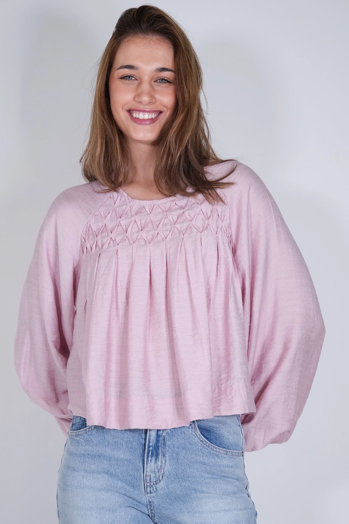 A smiling young woman wearing a light pink long-sleeve blouse with smocked detailing across the chest and light blue jeans, standing against a plain light gray studio background.