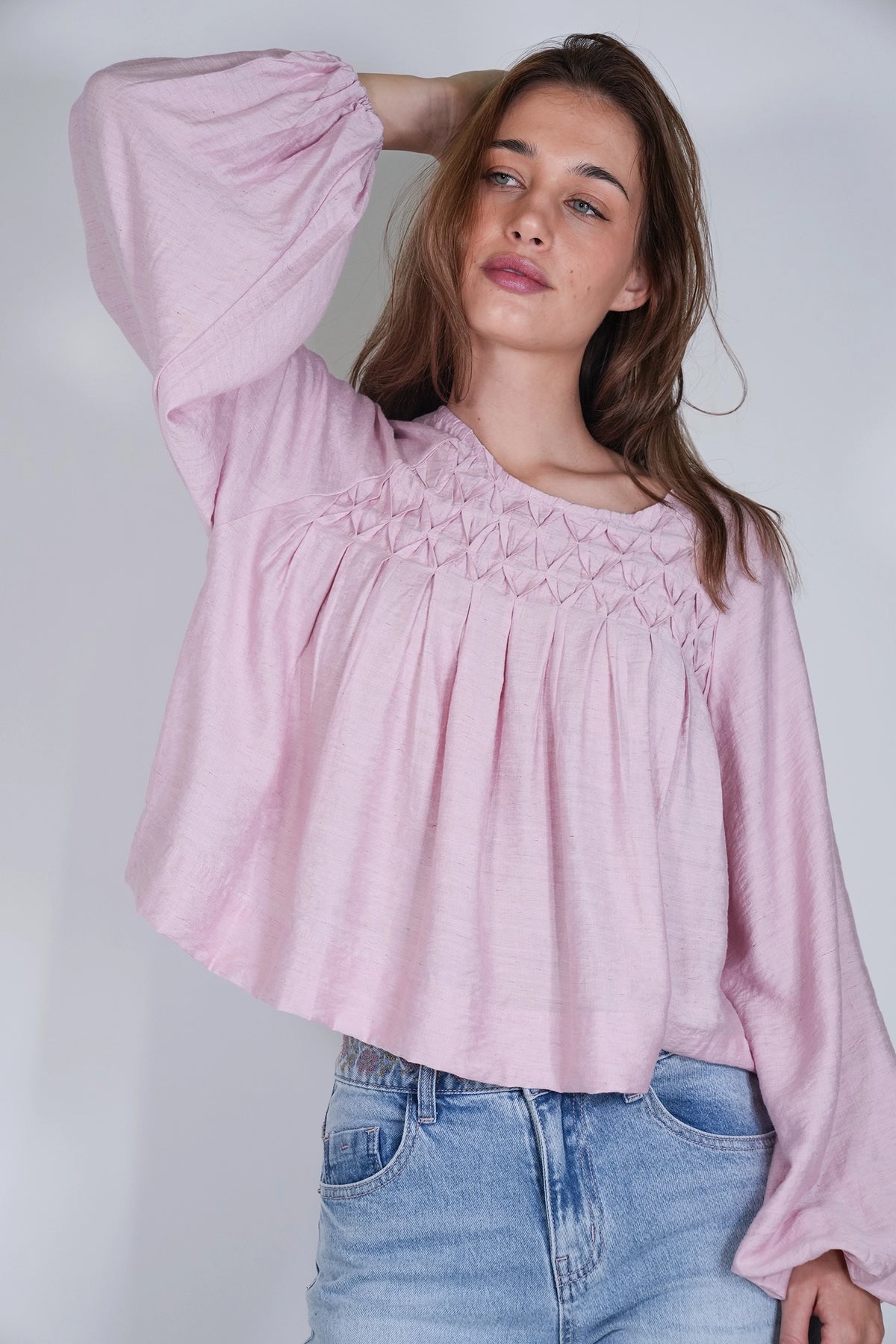 A woman wearing a light pink long-sleeve blouse with a smocked yoke and blue jeans, posing against a light gray studio background.