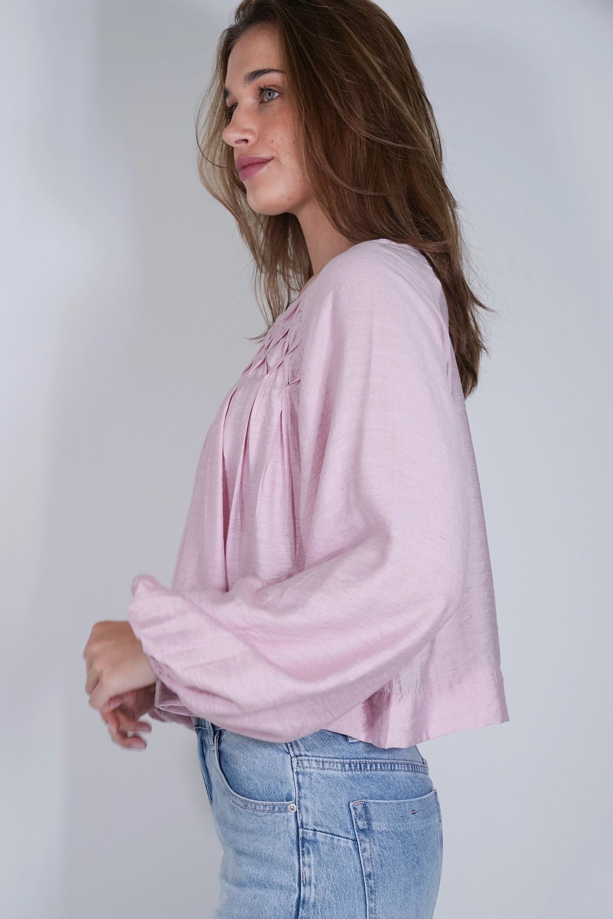 A woman wearing a light pink long-sleeve blouse with gathered detailing at the neckline and light blue jeans, shown from a side profile against a plain light gray background.