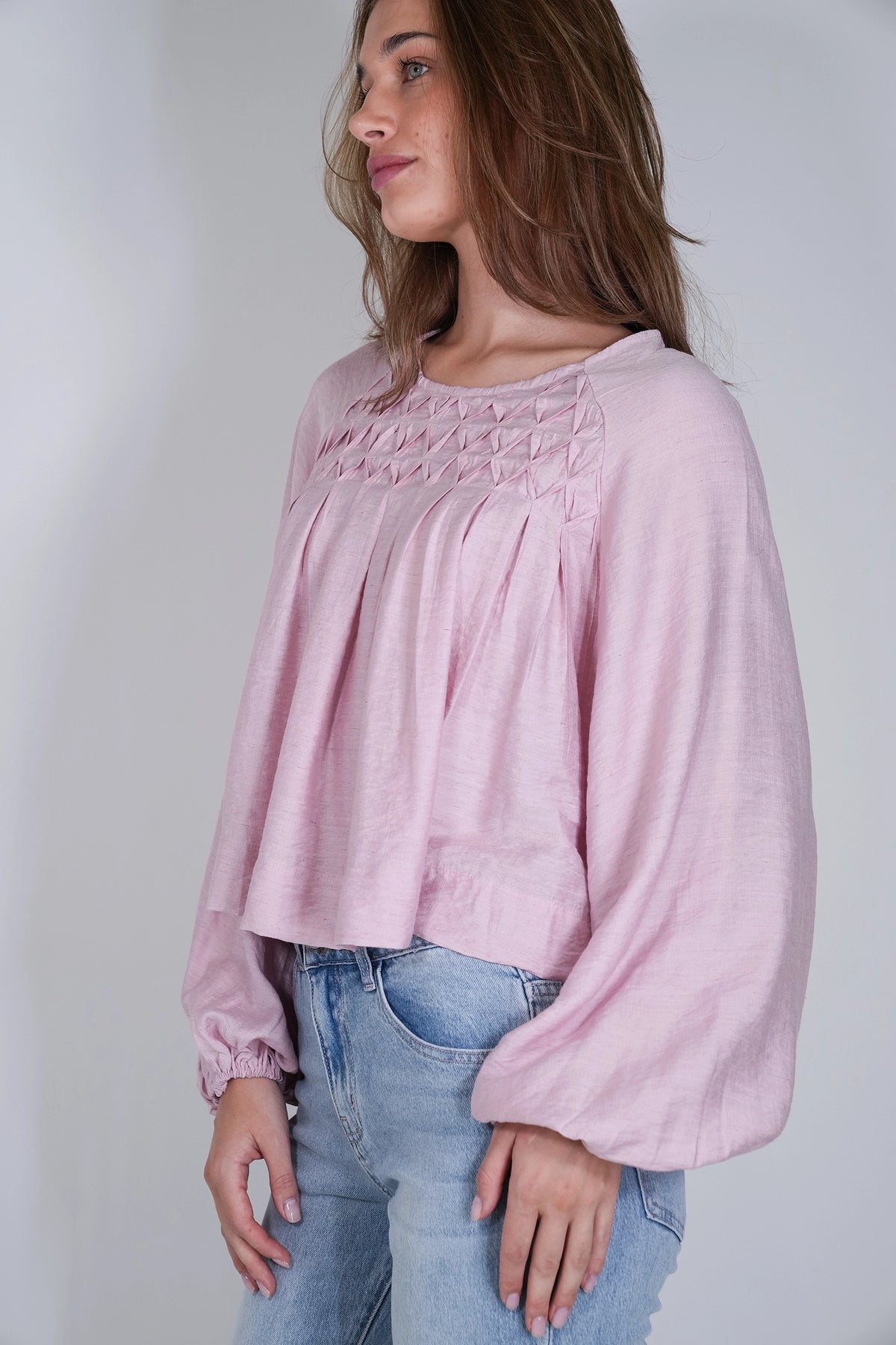 A woman wearing a light pink long-sleeve blouse with smocked detailing across the chest and gathered cuffs, paired with light blue jeans, photographed against a plain light gray background.