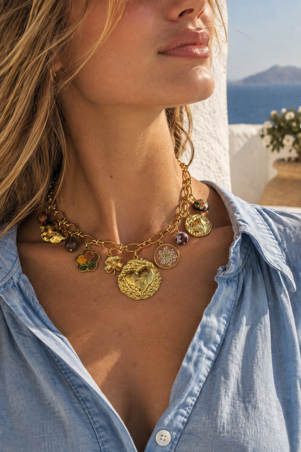 A close-up lifestyle shot of a woman wearing a gold-toned charm necklace with various colorful glass beads and medallion pendants, paired with a light blue denim shirt against a sunny outdoor background.