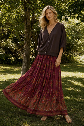 A woman standing outdoors in a grassy area with trees, wearing a dark brown button-front cardigan and a long pleated maxi skirt in deep burgundy with orange and gold paisley embroidery.