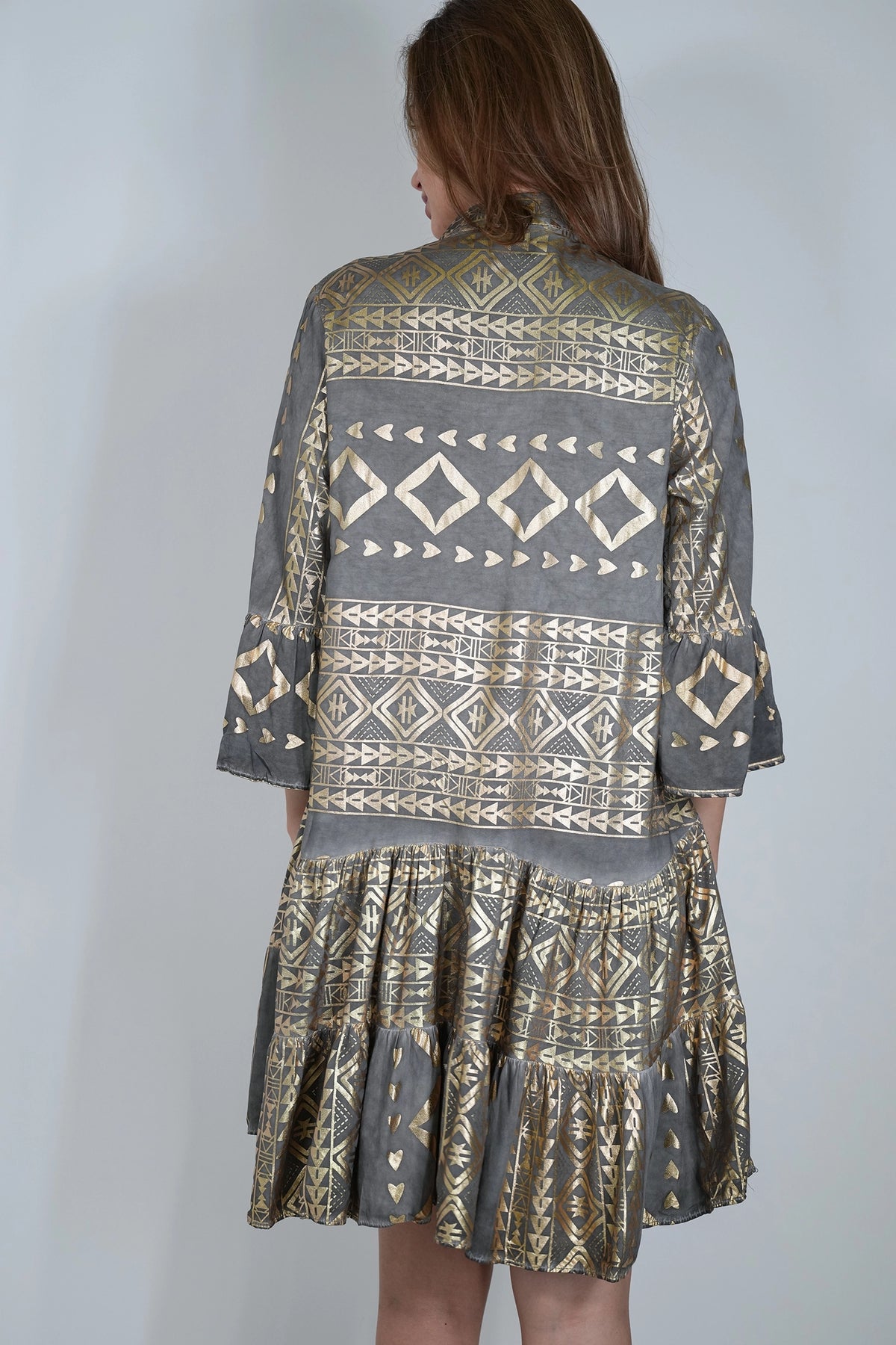 A woman viewed from the back wearing a gray and gold patterned dress with geometric tribal-style prints, set against a plain light gray background.