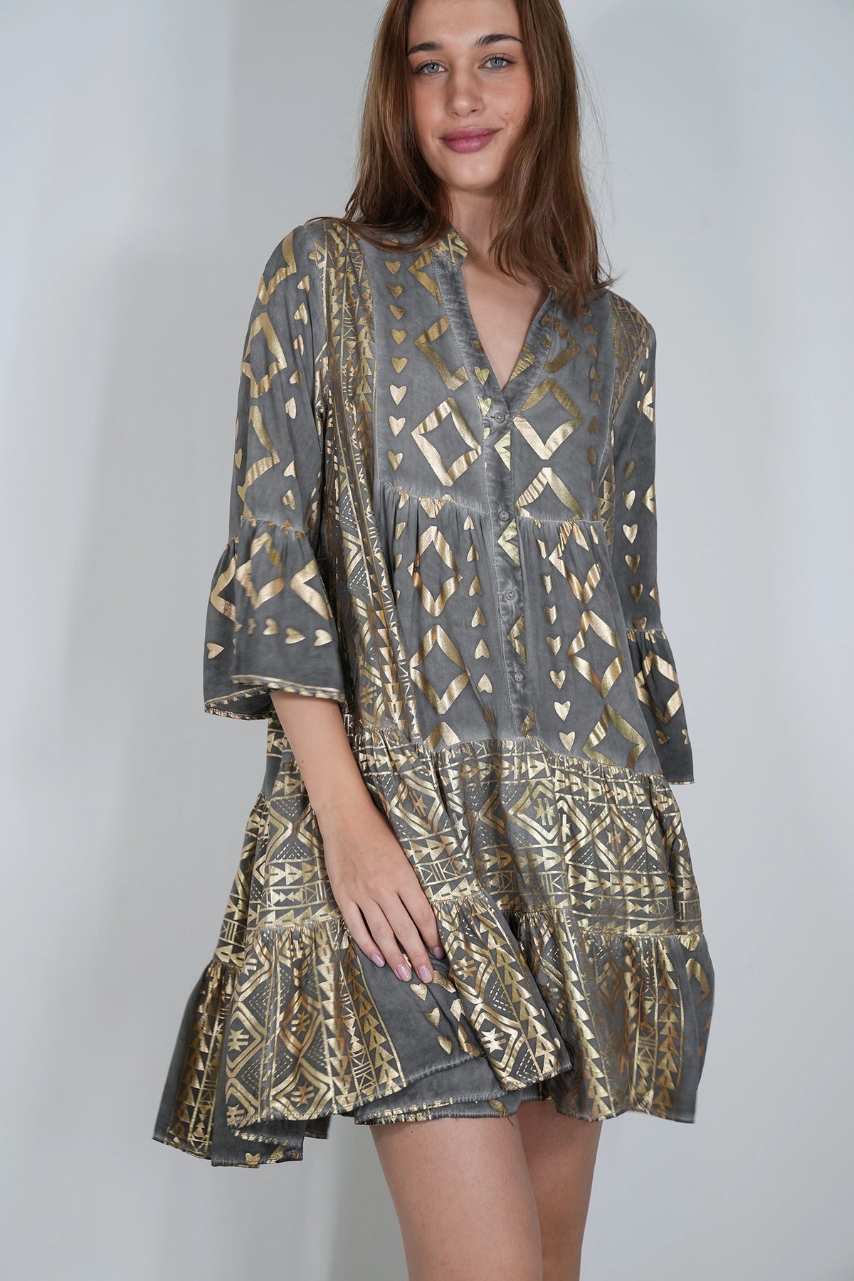 A woman wearing a gray and gold patterned bohemian-style tunic dress with bell sleeves, shown against a plain light gray studio background.