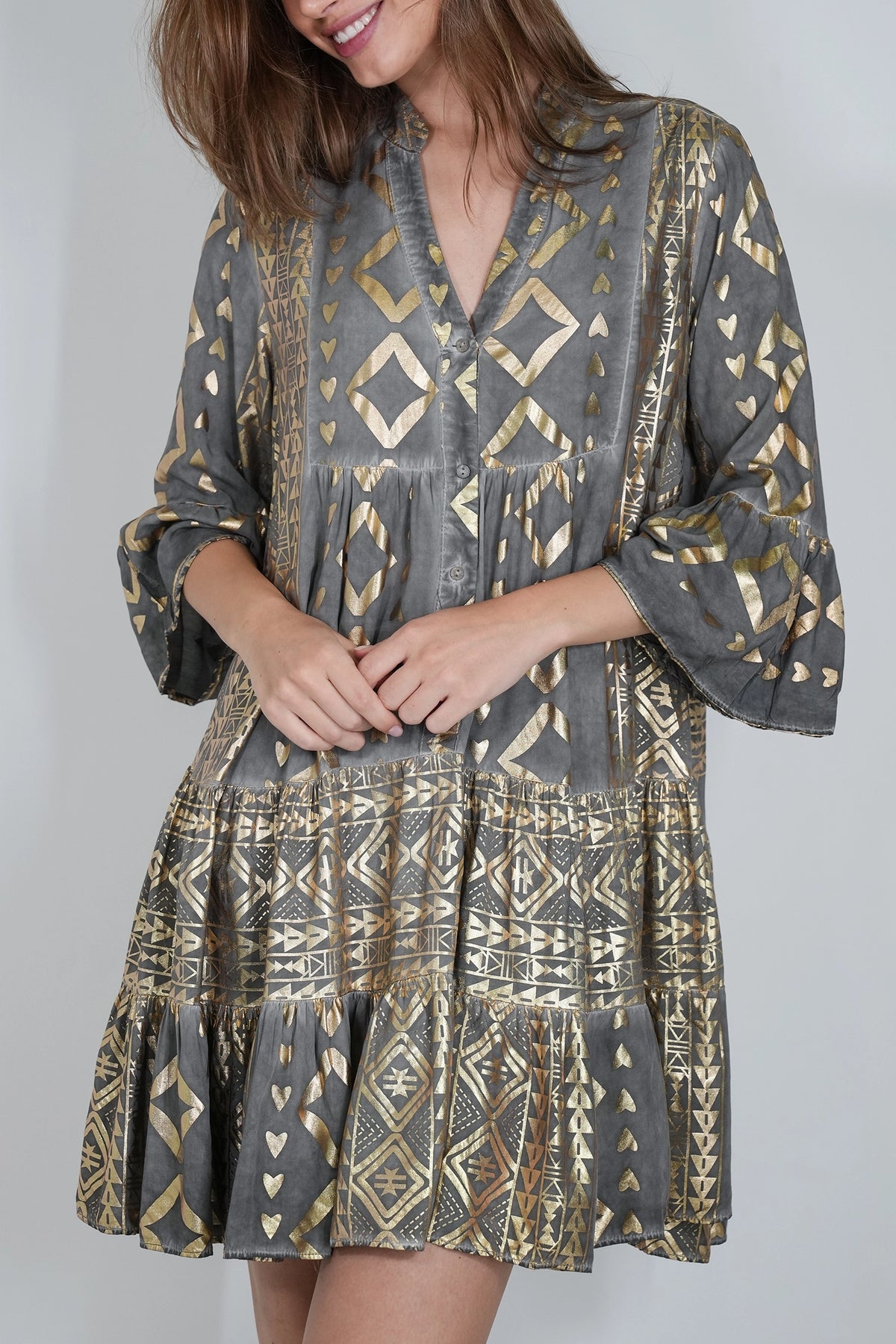A woman wearing a gray bohemian-style mini dress with gold geometric patterns, featuring a V-neckline, button-up front, and tiered skirt.