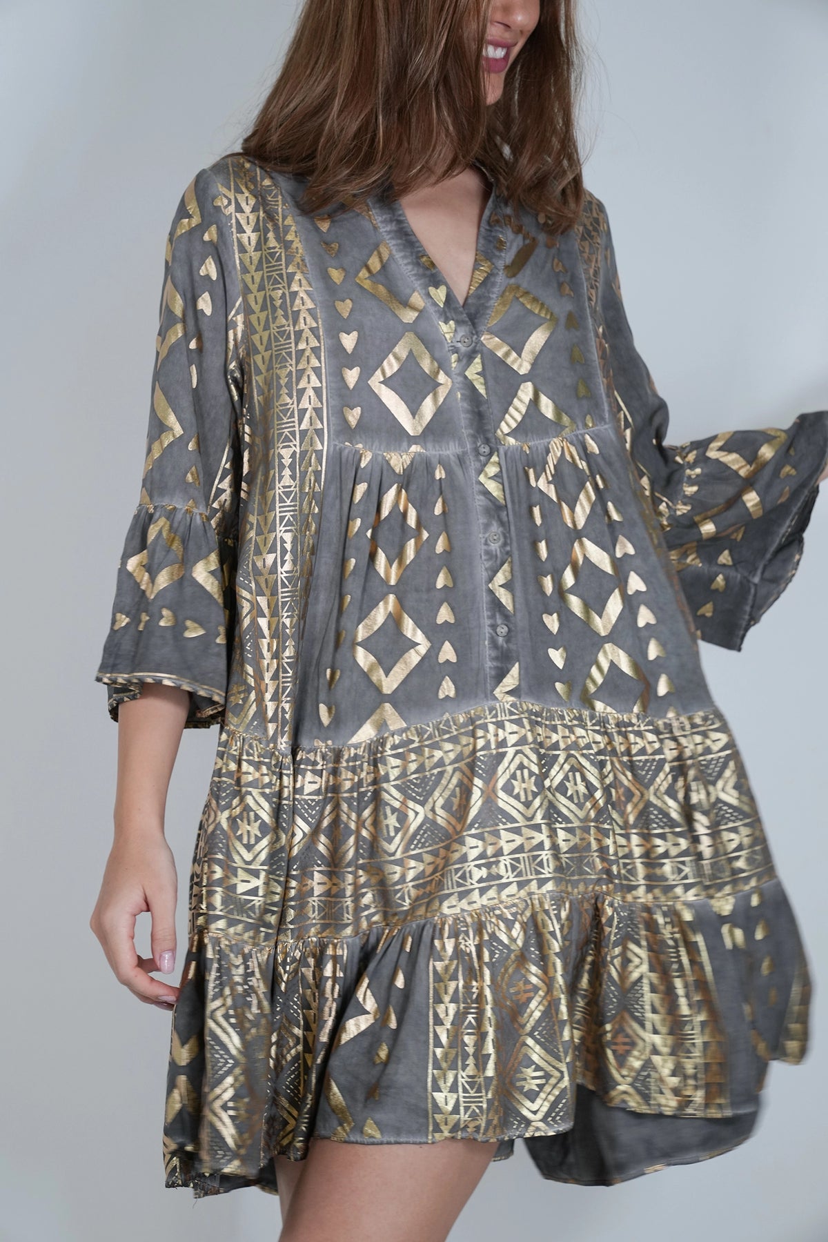 A woman wearing a gray and gold patterned tiered tunic dress with three-quarter bell sleeves, shown against a plain light gray background.
