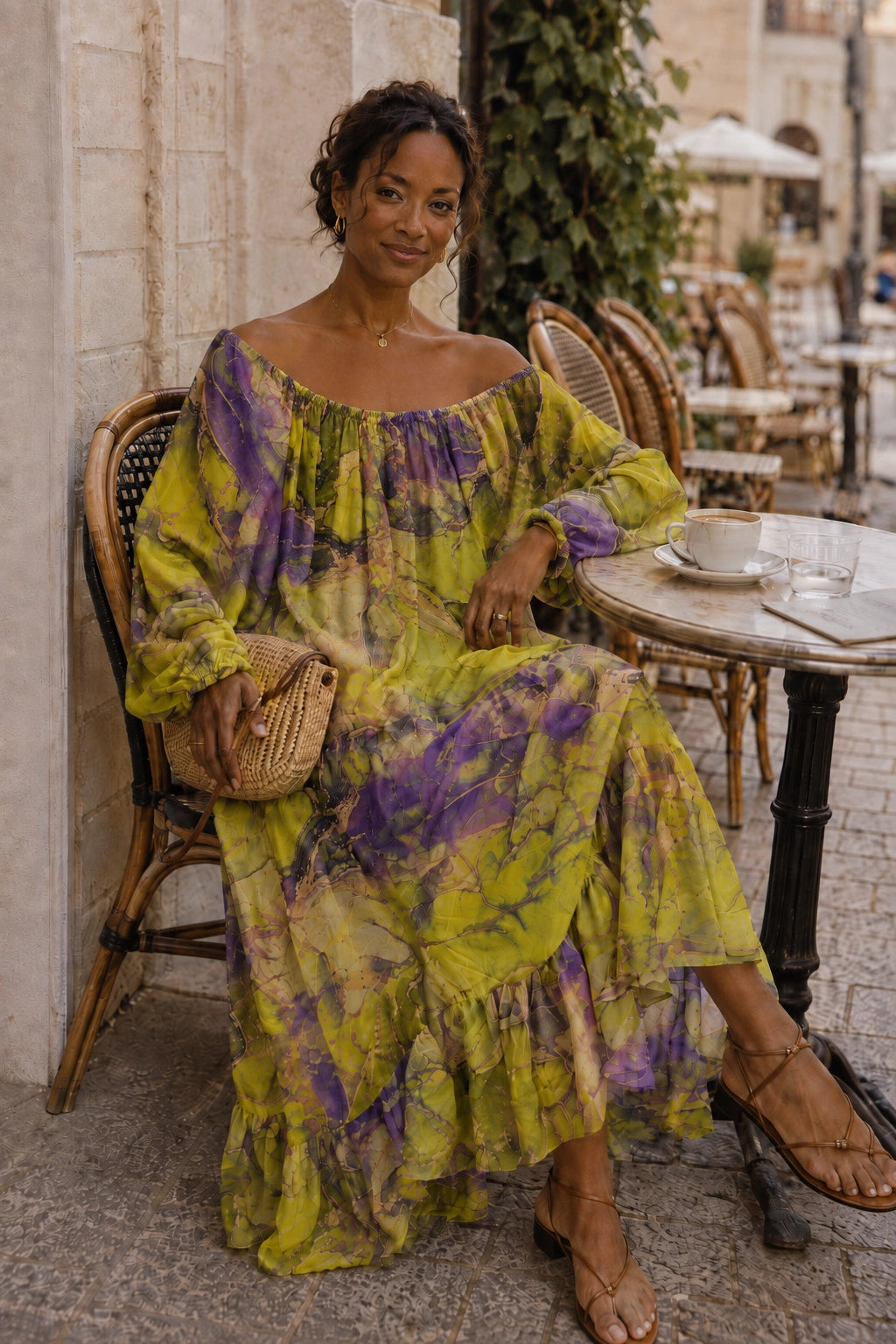 A woman sits on a wicker chair at an outdoor cafe wearing a long-sleeved, off-the-shoulder maxi dress with a yellow and purple tie-dye pattern. She is holding a small woven clutch bag.
