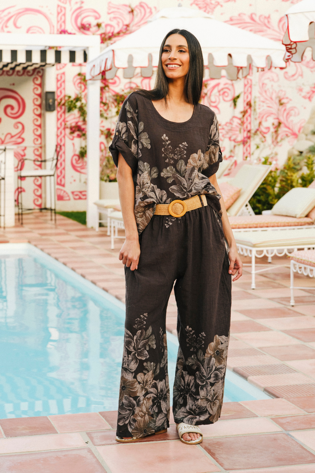 A woman stands by a swimming pool wearing a dark brown short-sleeve jumpsuit with a beige and gray floral print, styled with a wide tan belt. The background features a pink and white patterned wall, white patio furniture, and a tiled pool deck.