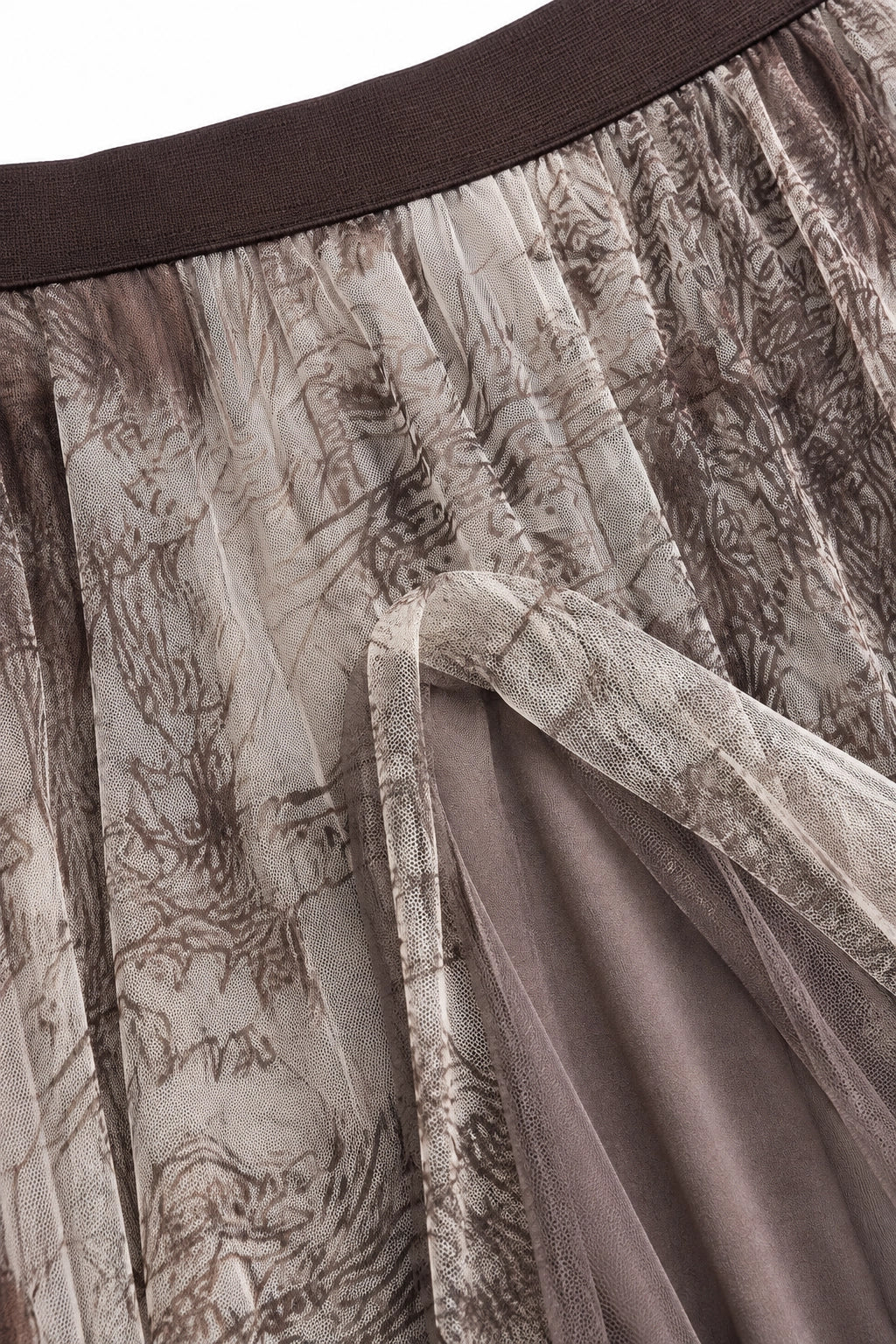 Close-up of a pleated skirt with a brown and beige abstract print, featuring a solid dark brown waistband and a sheer tulle overlay.