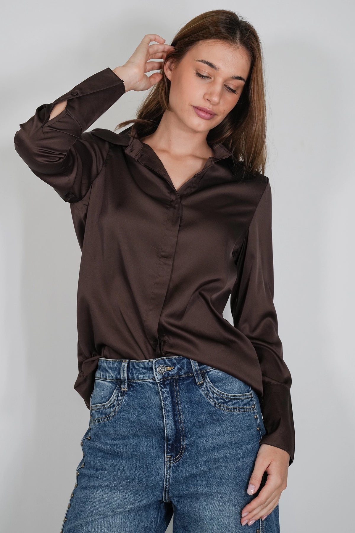 A woman wearing a dark brown satin long-sleeve button-up shirt and blue jeans, posing against a light gray studio background.