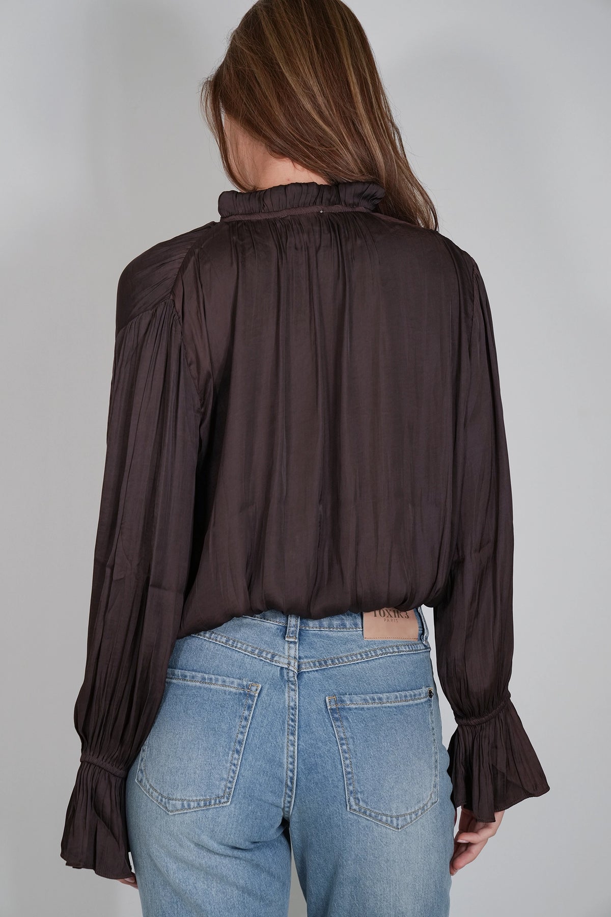 A woman viewed from the back wearing a dark brown long-sleeve blouse with a gathered high neck and flared ruffled cuffs, paired with light blue jeans, against a plain light gray studio background.