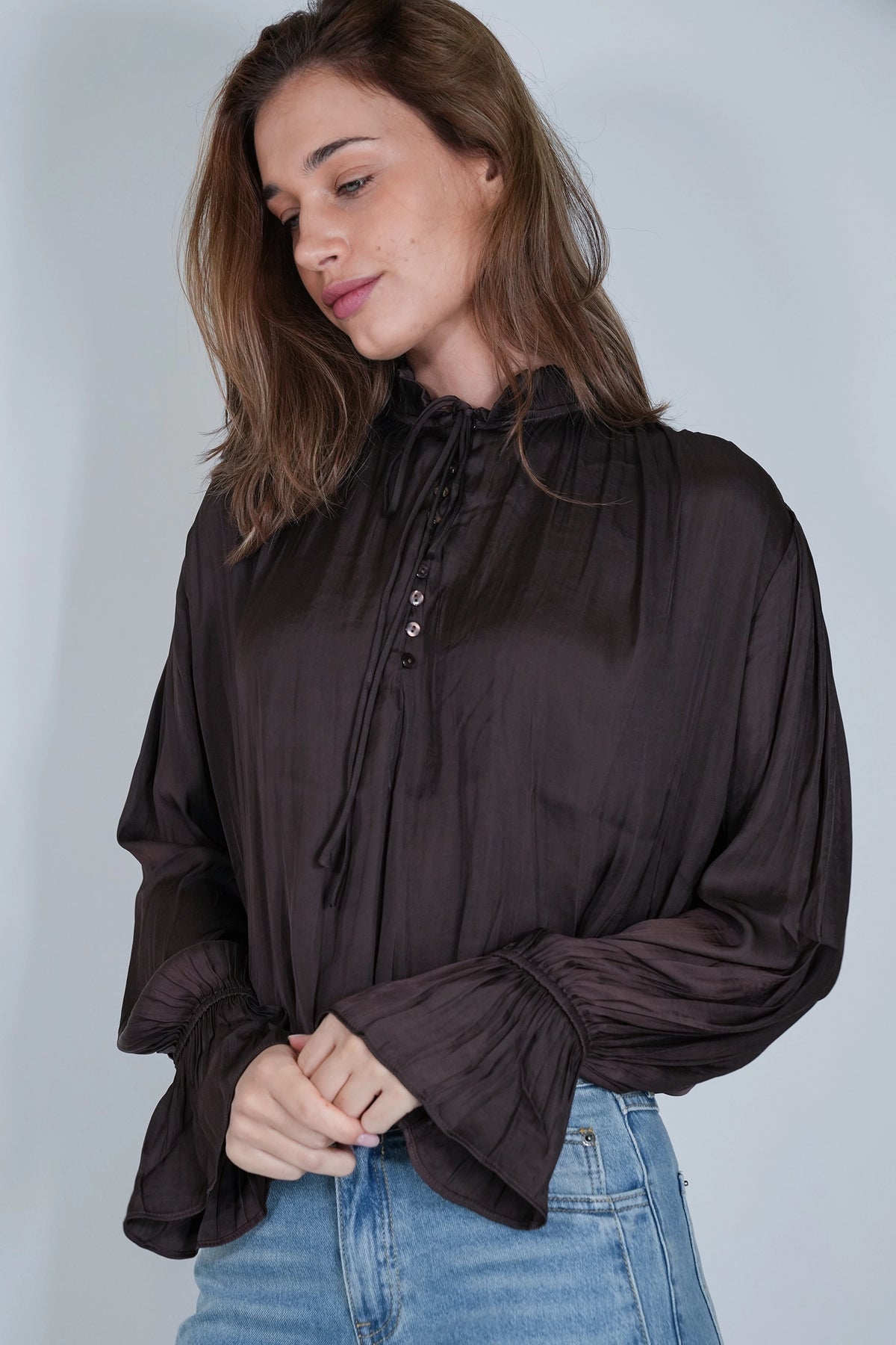 A woman wearing a dark brown sheer long-sleeve blouse with a high neck, front tie detail, and gathered cuffs, paired with blue jeans, against a light gray studio background.