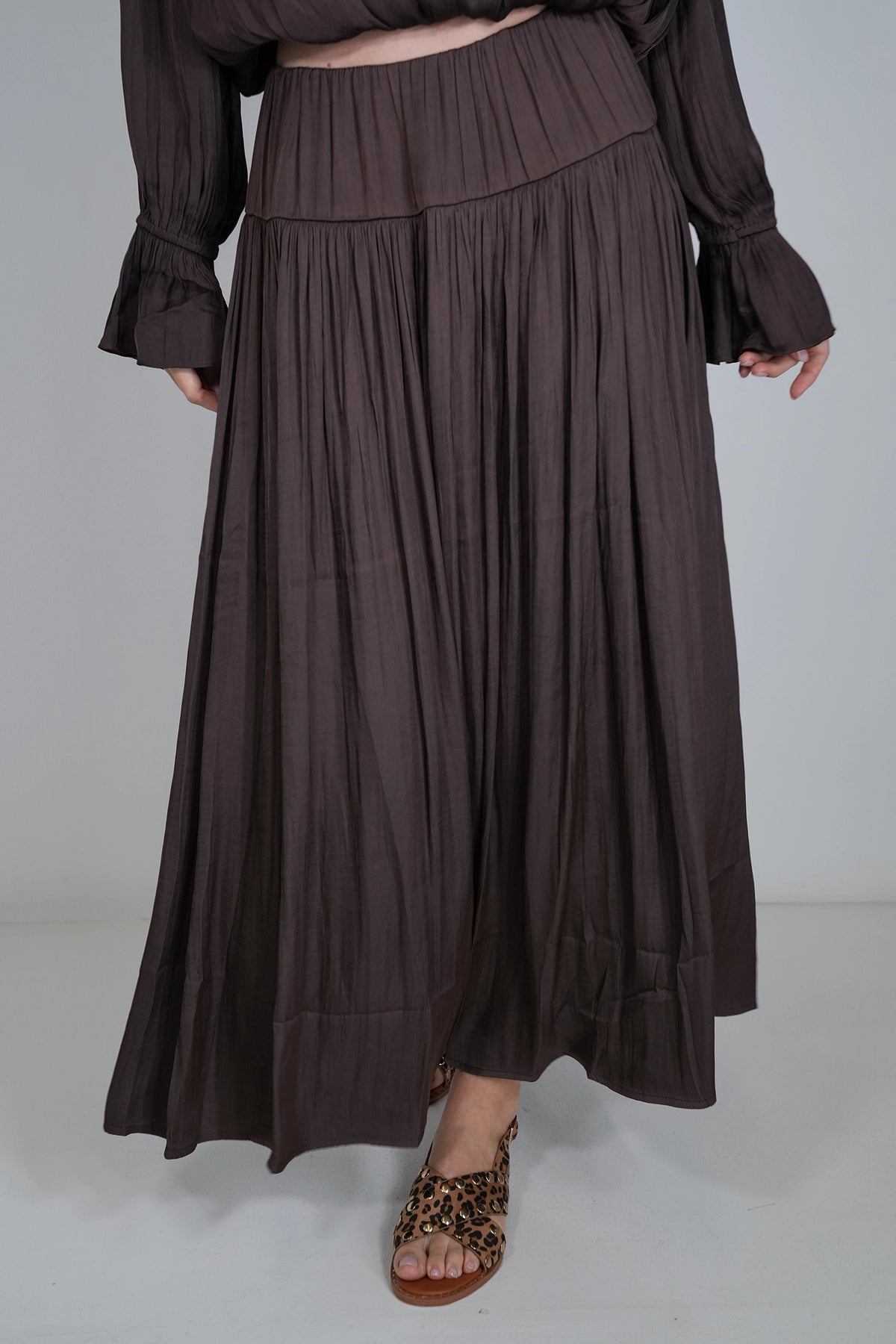 A dark brown pleated maxi skirt worn by a model, paired with leopard print flat sandals, shown against a plain light gray studio background.