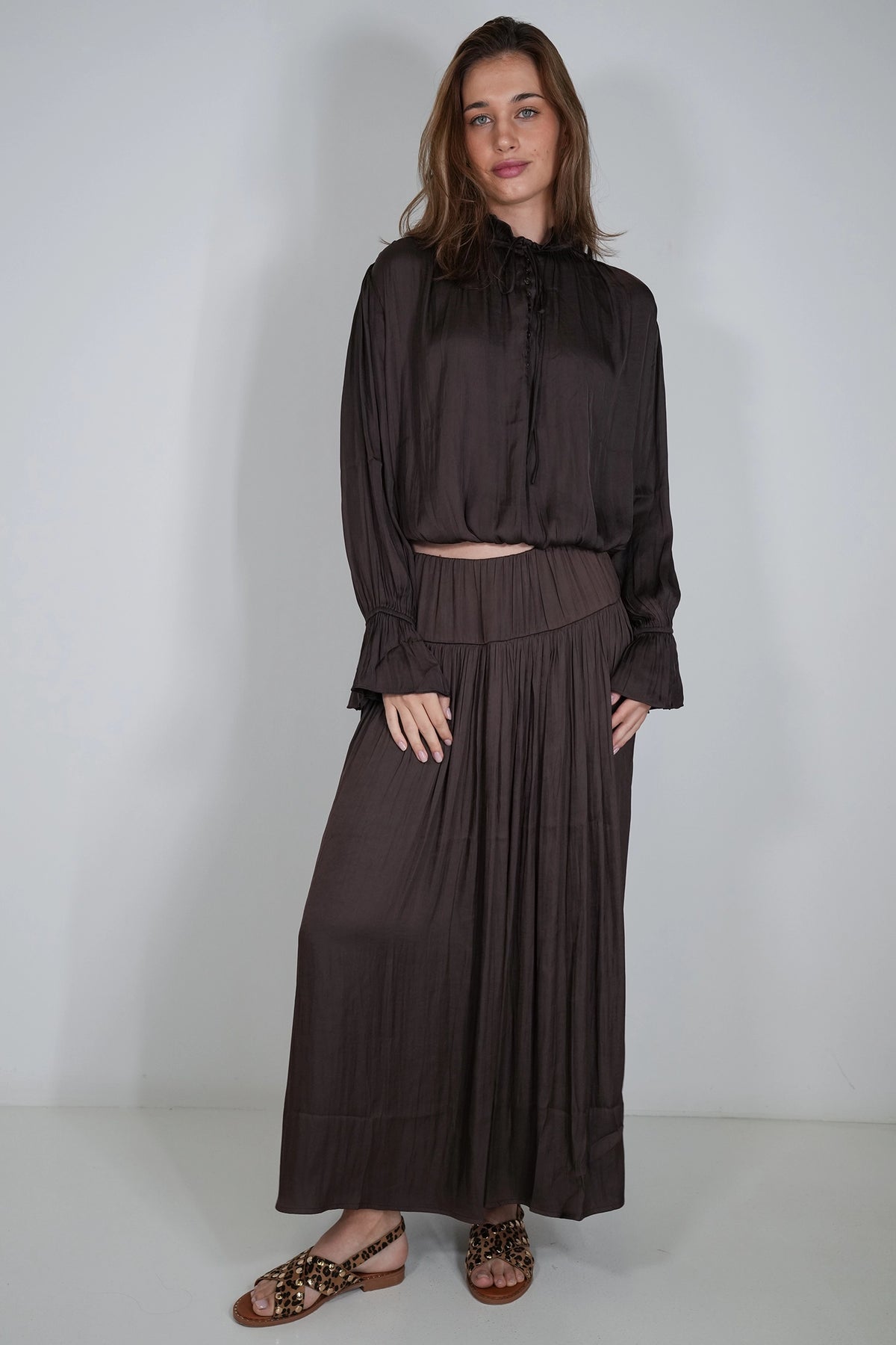 A woman modeling a dark brown two-piece set consisting of a long-sleeve high-neck blouse and a matching pleated maxi skirt, paired with leopard-print flat sandals, against a plain light gray studio background.