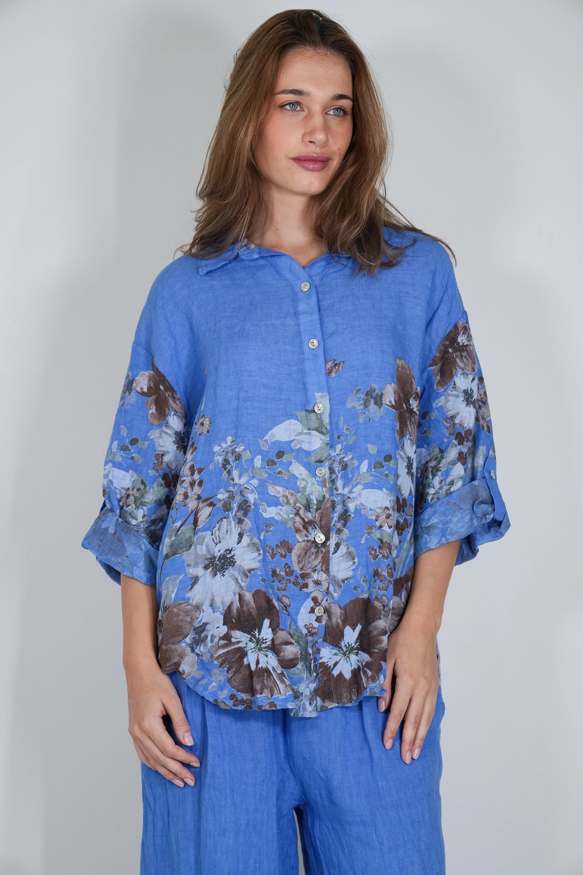 A woman modeling a blue button-up shirt with a brown and white floral print, paired with matching blue pants, against a plain light gray background.