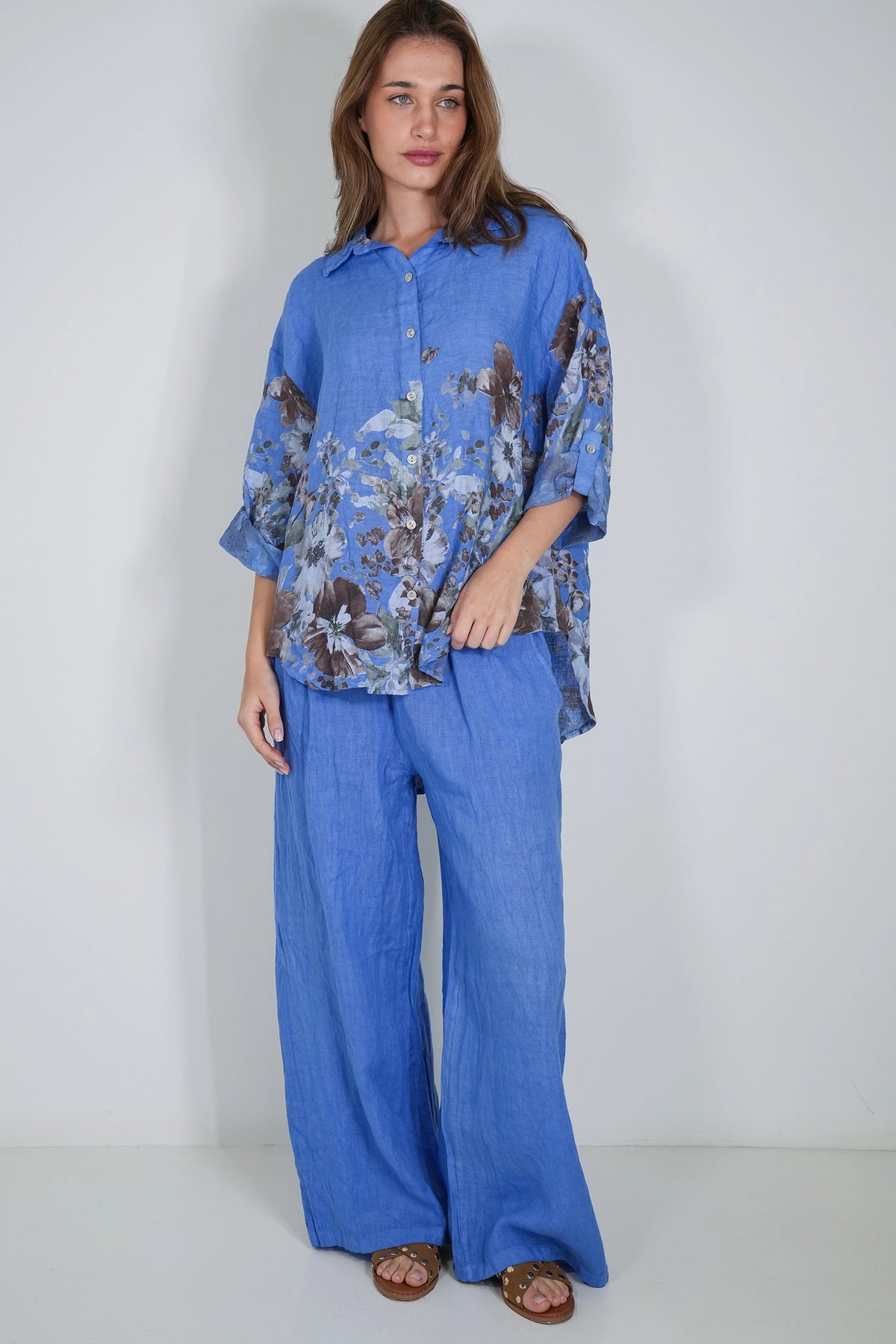 A woman modeling a blue two-piece linen set consisting of a button-down shirt with a brown floral print and matching wide-leg pants, standing against a plain white studio background.