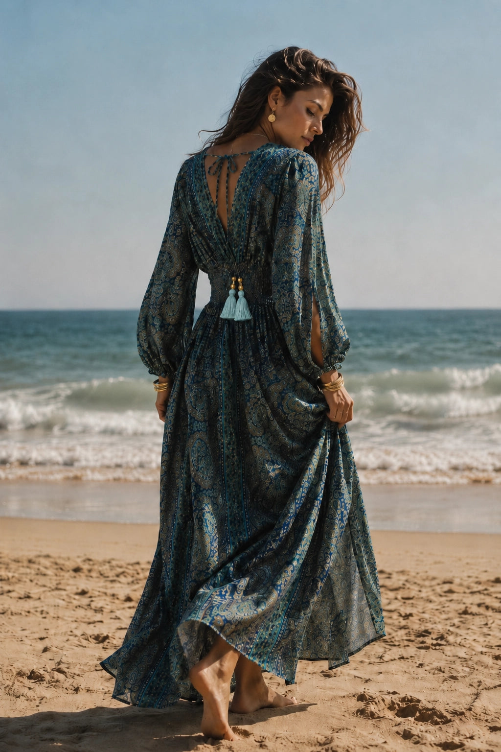 A woman stands on a sandy beach facing away from the camera, wearing a long-sleeved, open-back maxi dress with a blue and green bohemian pattern and tassel details.