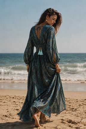 A woman stands on a sandy beach facing away from the camera, wearing a long-sleeved, open-back maxi dress with a blue and green bohemian pattern and tassel details.