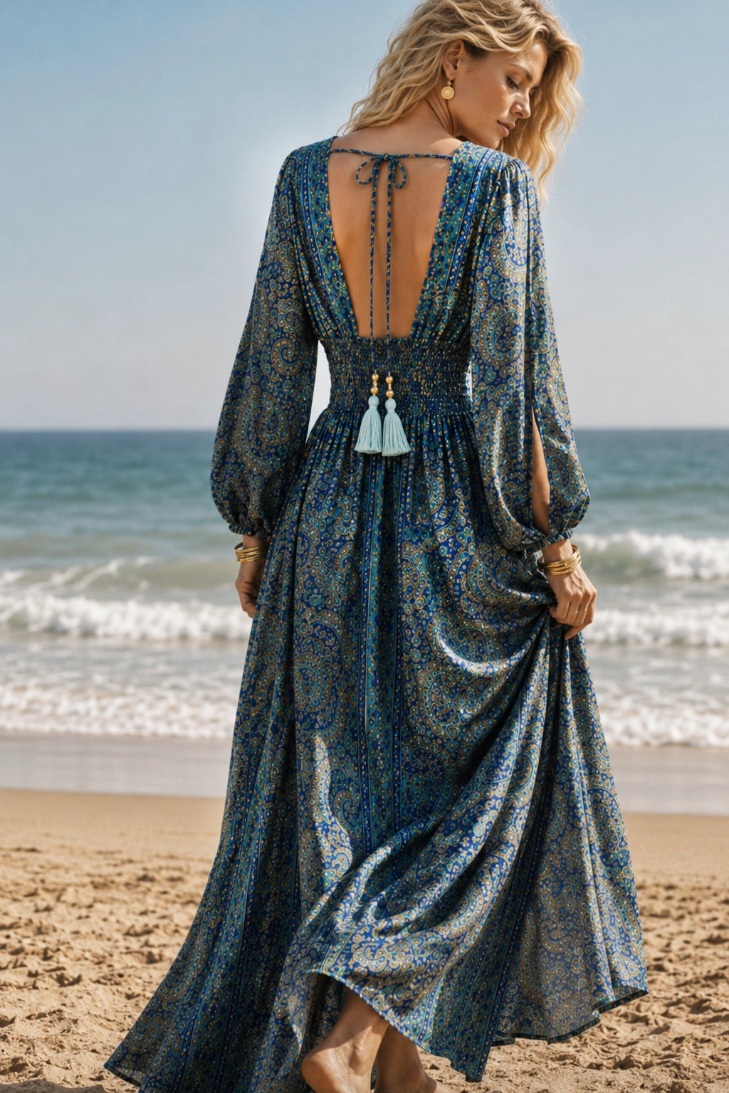 A woman on a beach wearing a long-sleeve maxi dress with a deep open back, tie detail, and tassel accents. The dress has a blue and gray paisley pattern and is photographed from behind.