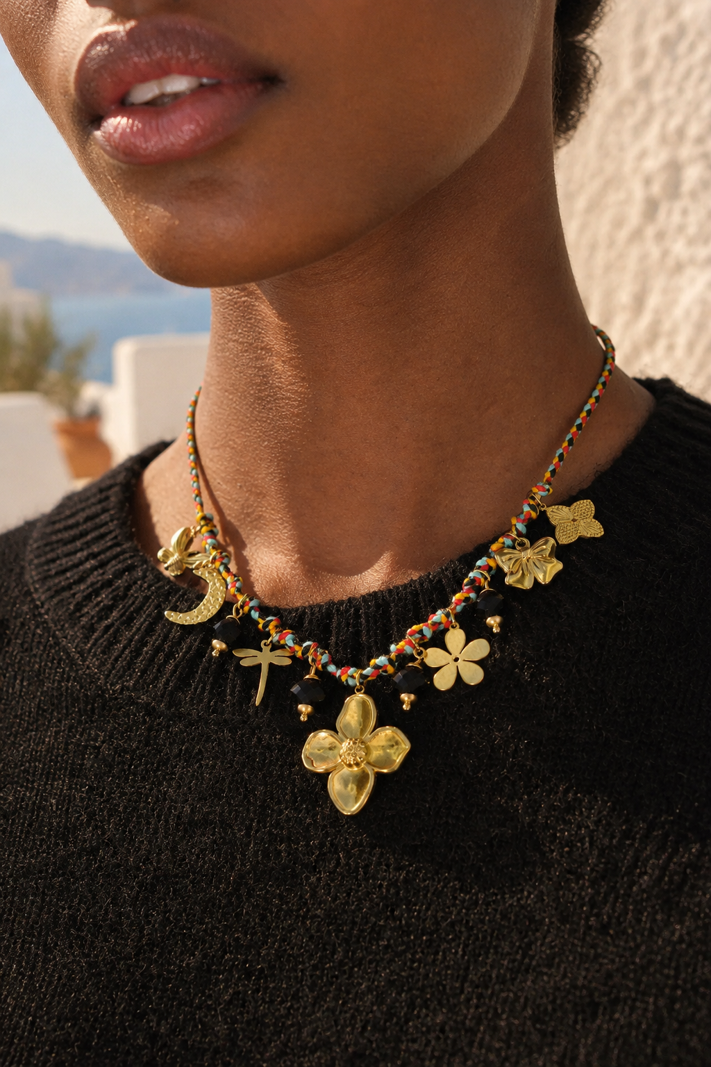 A close-up of a model wearing a gold-toned statement necklace with floral, crescent moon, and dragonfly charms, paired with a black knit sweater.