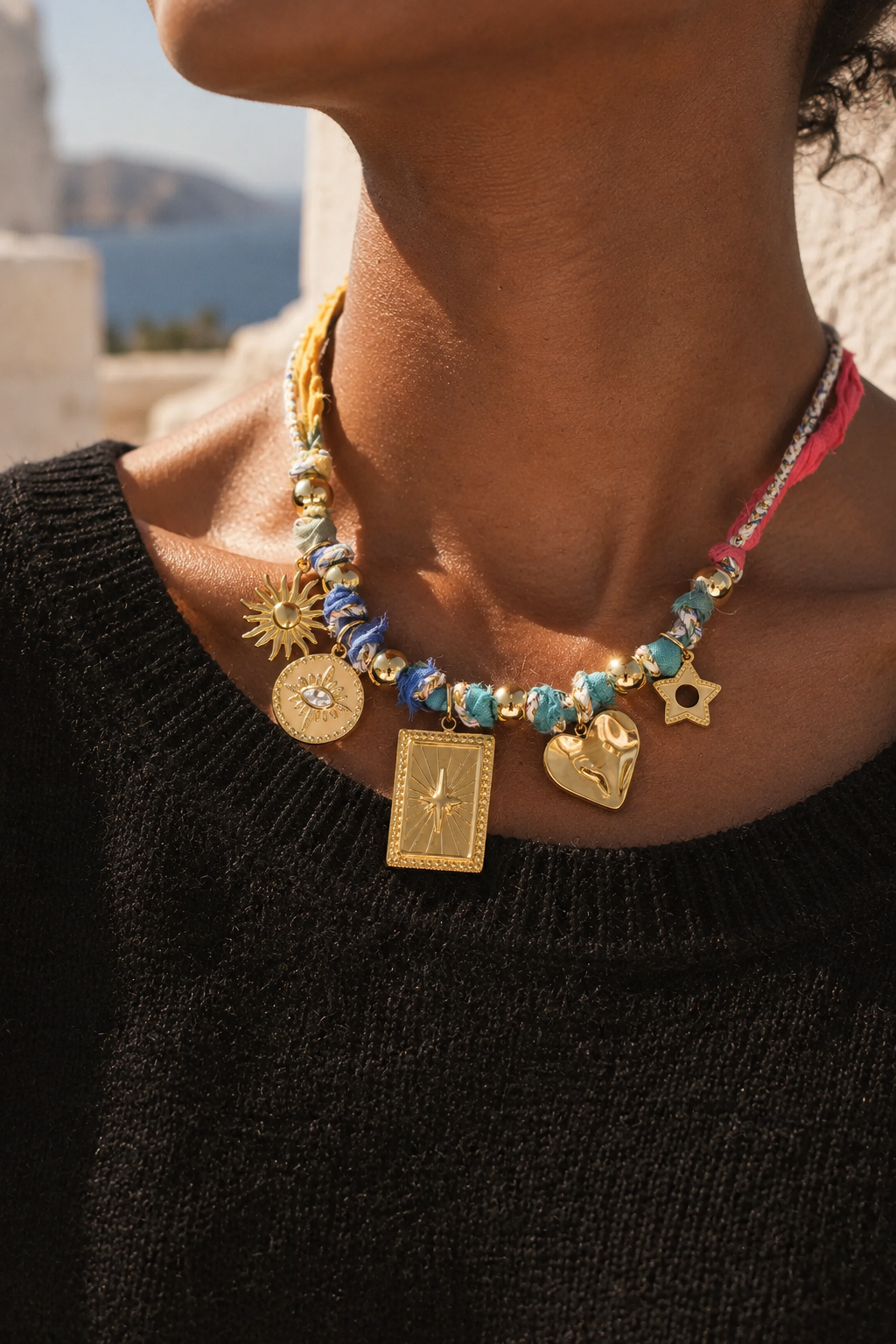 A close-up of a person wearing a bohemian-style beaded necklace with gold charms, including a card, sun, and heart, set against a warm outdoor background.