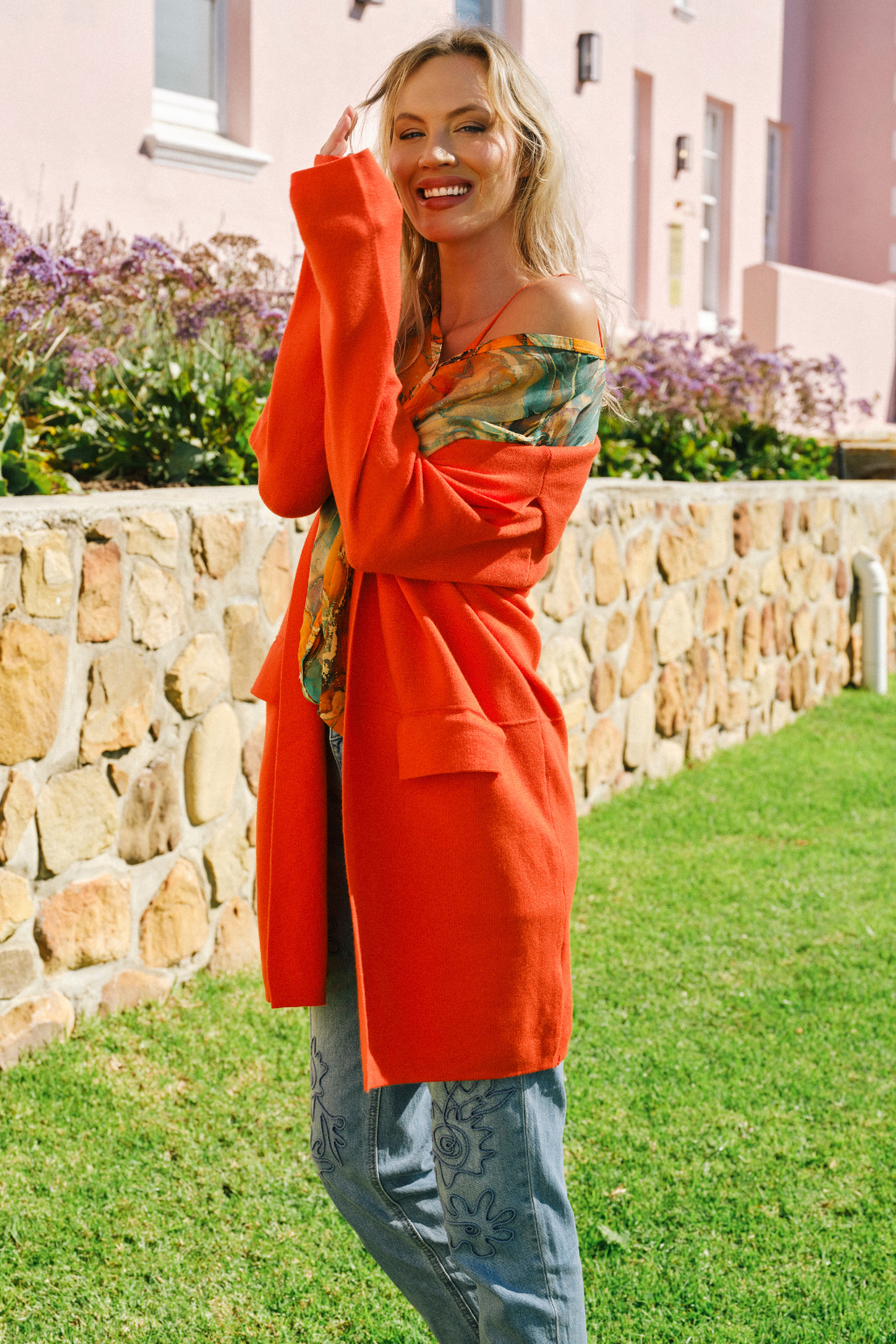 A woman posing outdoors in a vibrant orange oversized cardigan over a colorful off-the-shoulder top and blue jeans, set against a stone wall and green lawn.