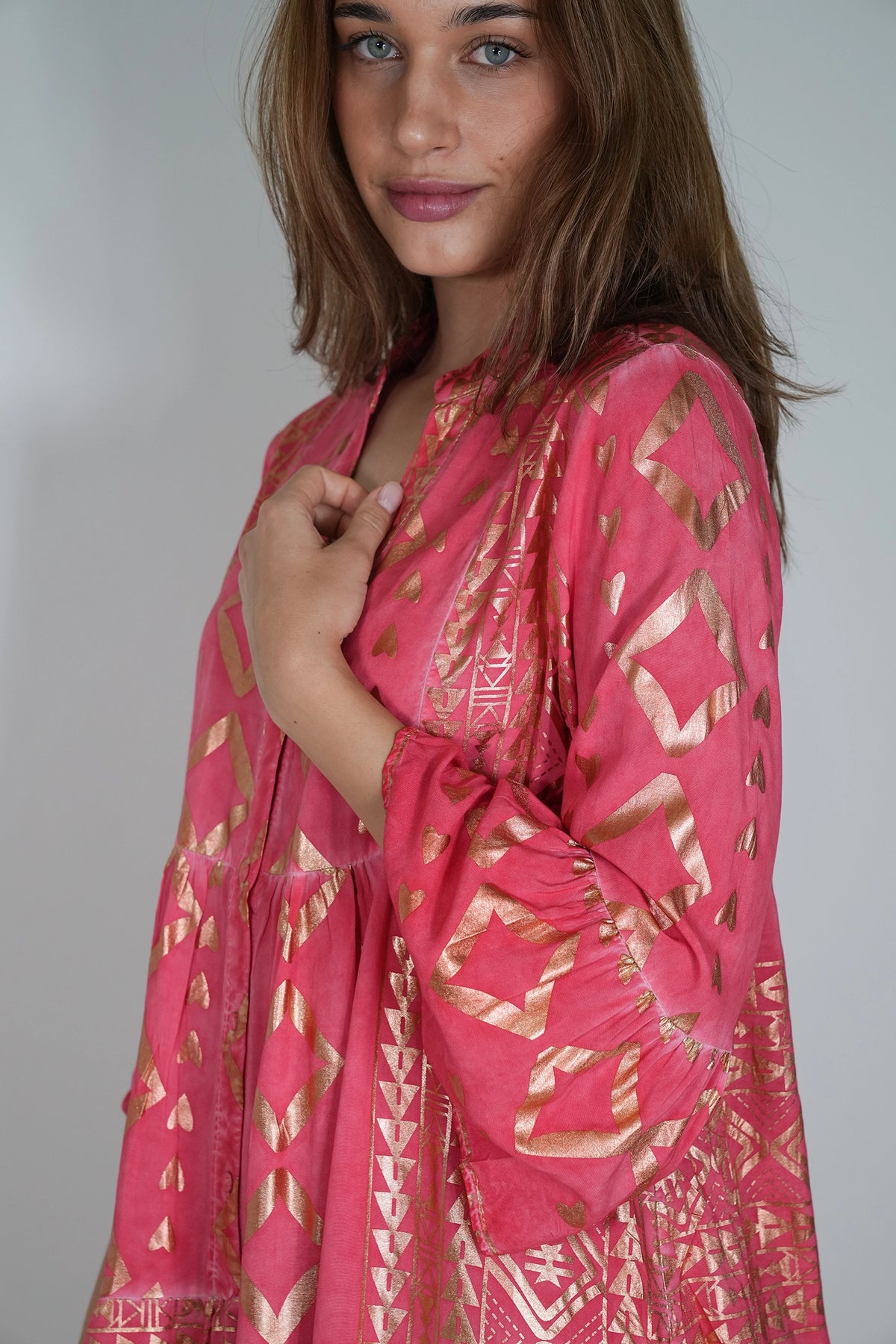 A woman wearing a pink cotton kaftan dress with a metallic gold geometric pattern, photographed against a neutral light gray background.