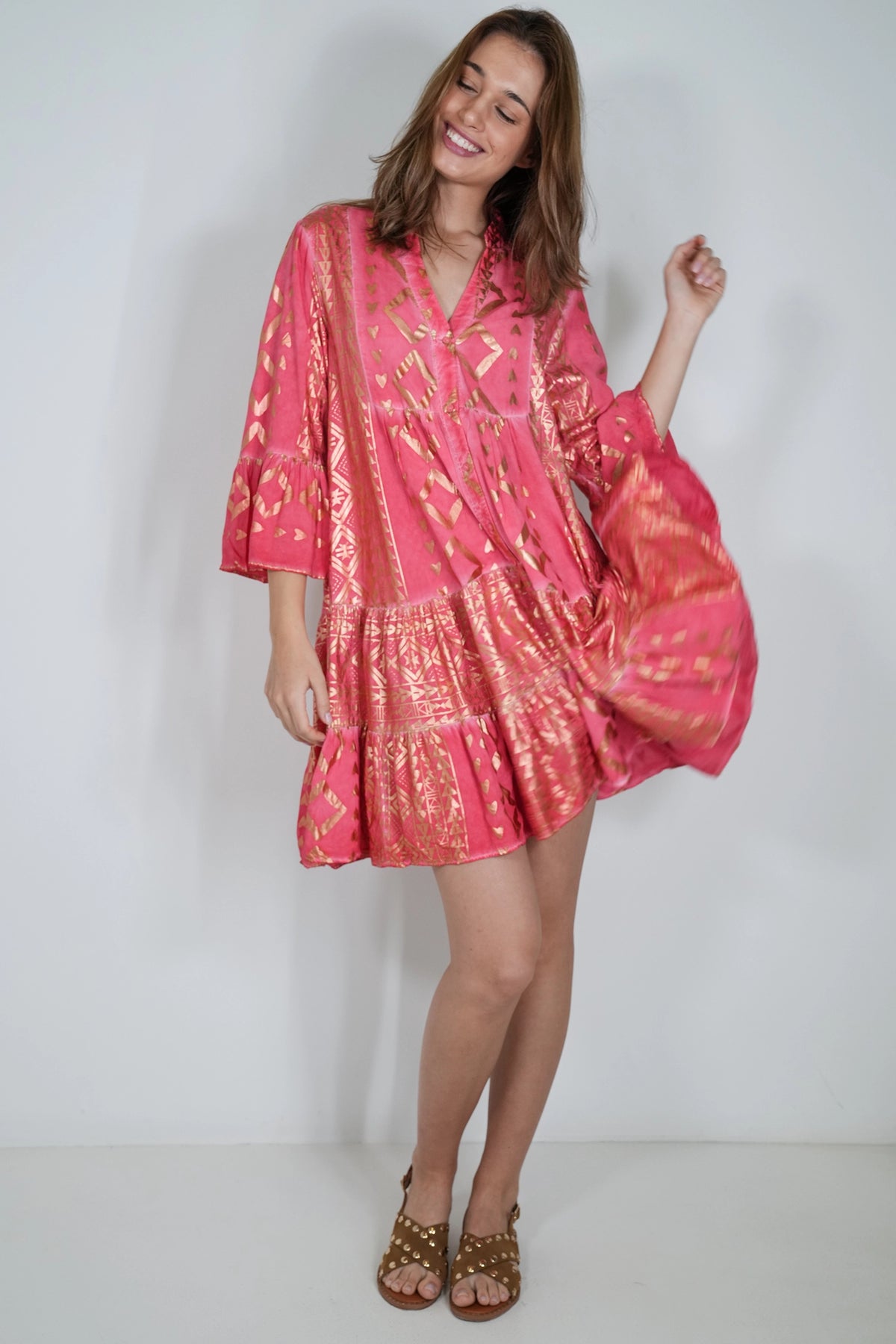 A woman modeling a pink kimono-style cover-up dress with gold patterns, standing against a plain white studio background.
