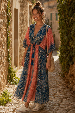 A woman stands on a cobblestone alleyway wearing a long, loose bohemian maxi dress with a blue and coral-orange paisley pattern and tassel tie. She is holding a woven straw bag.