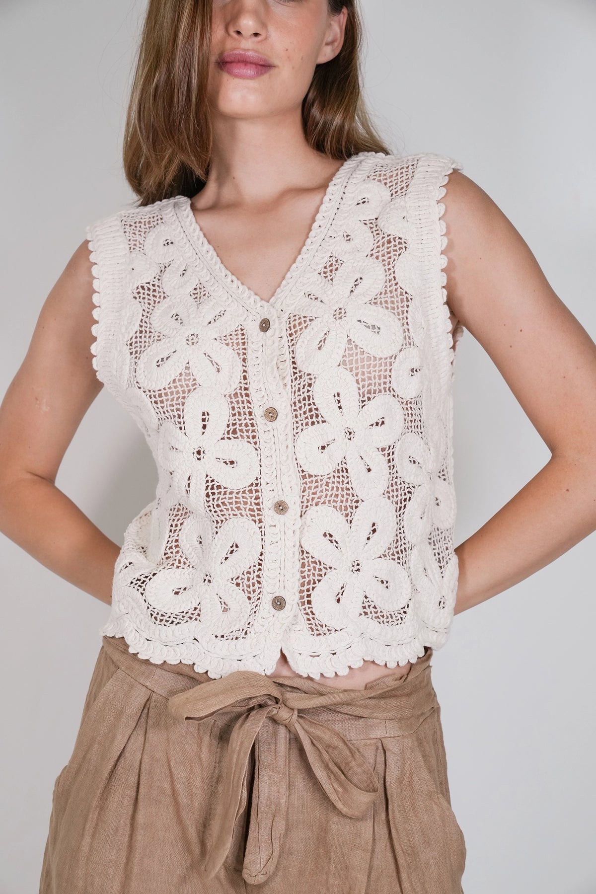 A woman wearing a sleeveless cream crochet vest with floral lace patterns and brown buttons, paired with tan pants, against a light gray background.