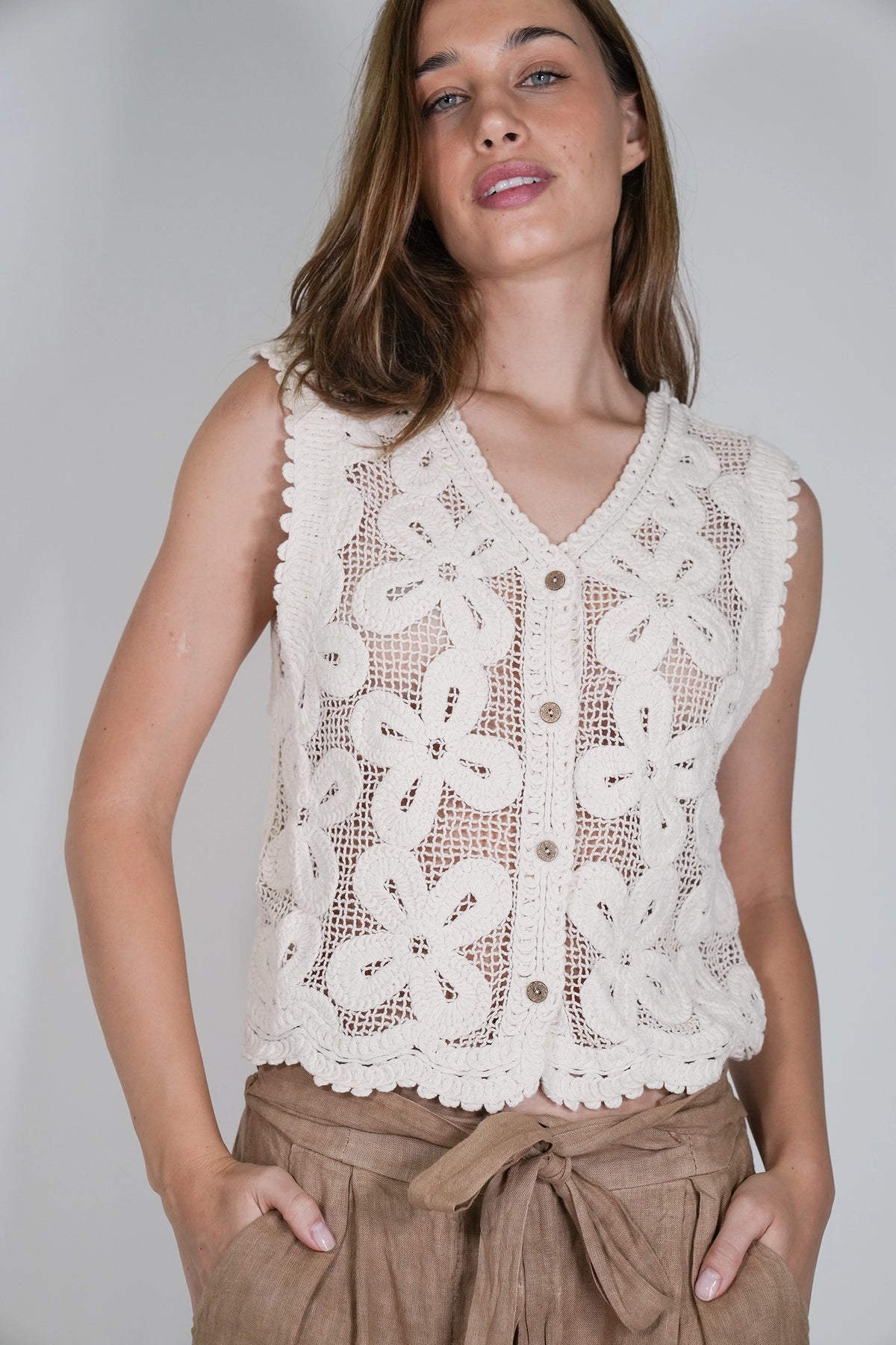 A woman wearing a sleeveless cream crochet vest with a button front over a brown top, paired with brown pants, against a plain light gray background.