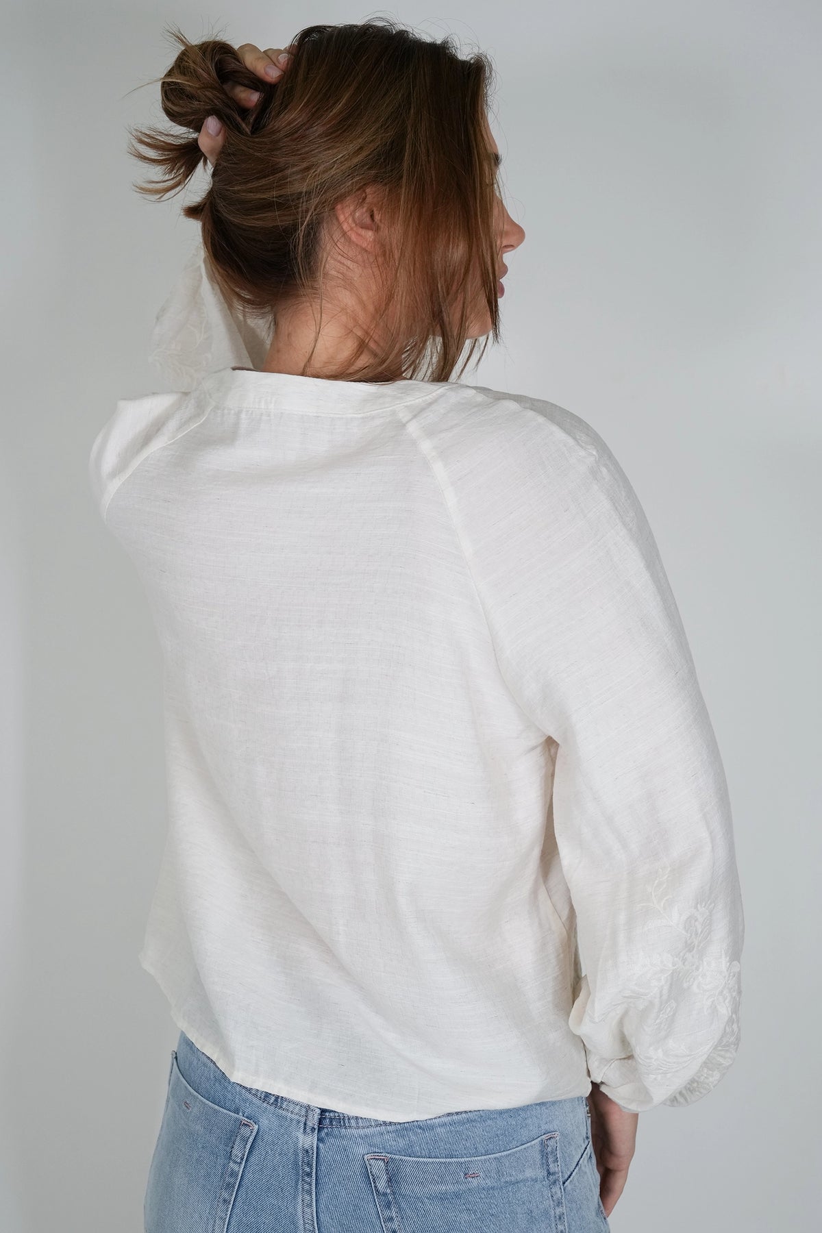A woman shown from the back wearing a white long-sleeve blouse and light blue jeans against a plain light gray background.