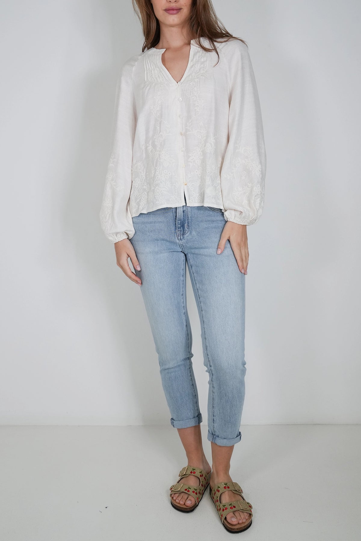 A woman modeling a white long-sleeve blouse with a split V-neck and light-wash blue cropped jeans, standing against a plain light gray studio background.