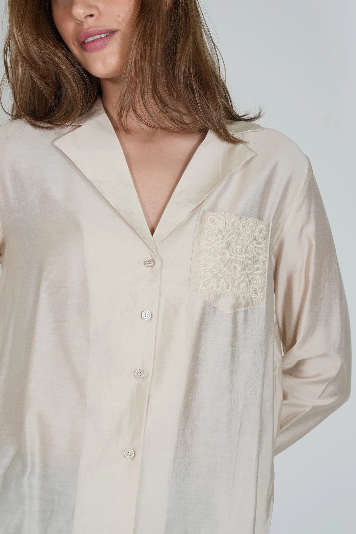 A close-up of a woman wearing a light beige long-sleeve button-front shirt with a pointed collar and a decorative embroidered pocket on the chest.