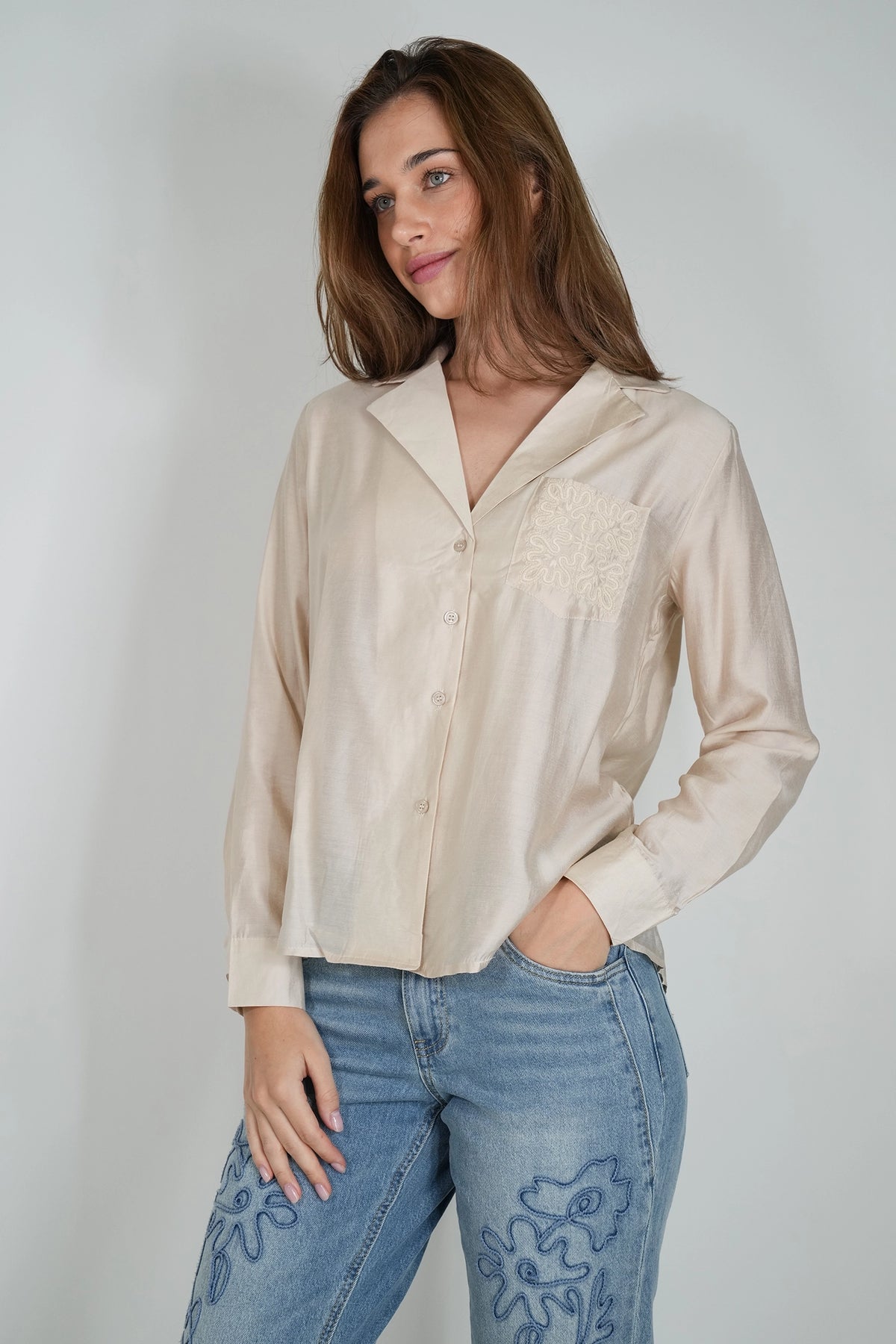 A woman wearing a light beige long-sleeve button-up shirt with a lace chest pocket detail, paired with blue jeans featuring blue floral embroidery, posed against a light gray studio background.