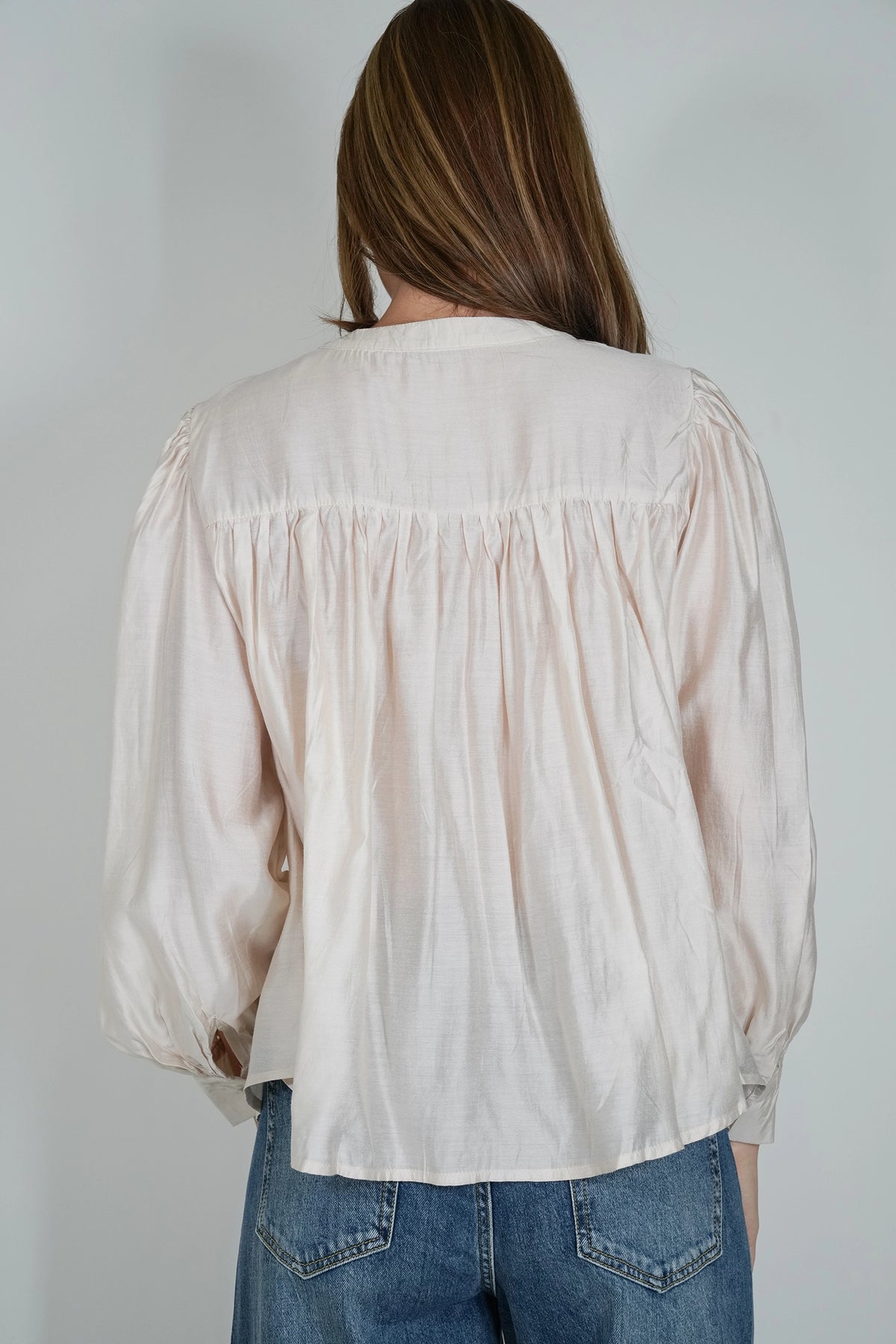 A woman viewed from behind wearing a light cream-colored long-sleeve blouse with a gathered, smocked back design and blue jeans, set against a plain light gray background.
